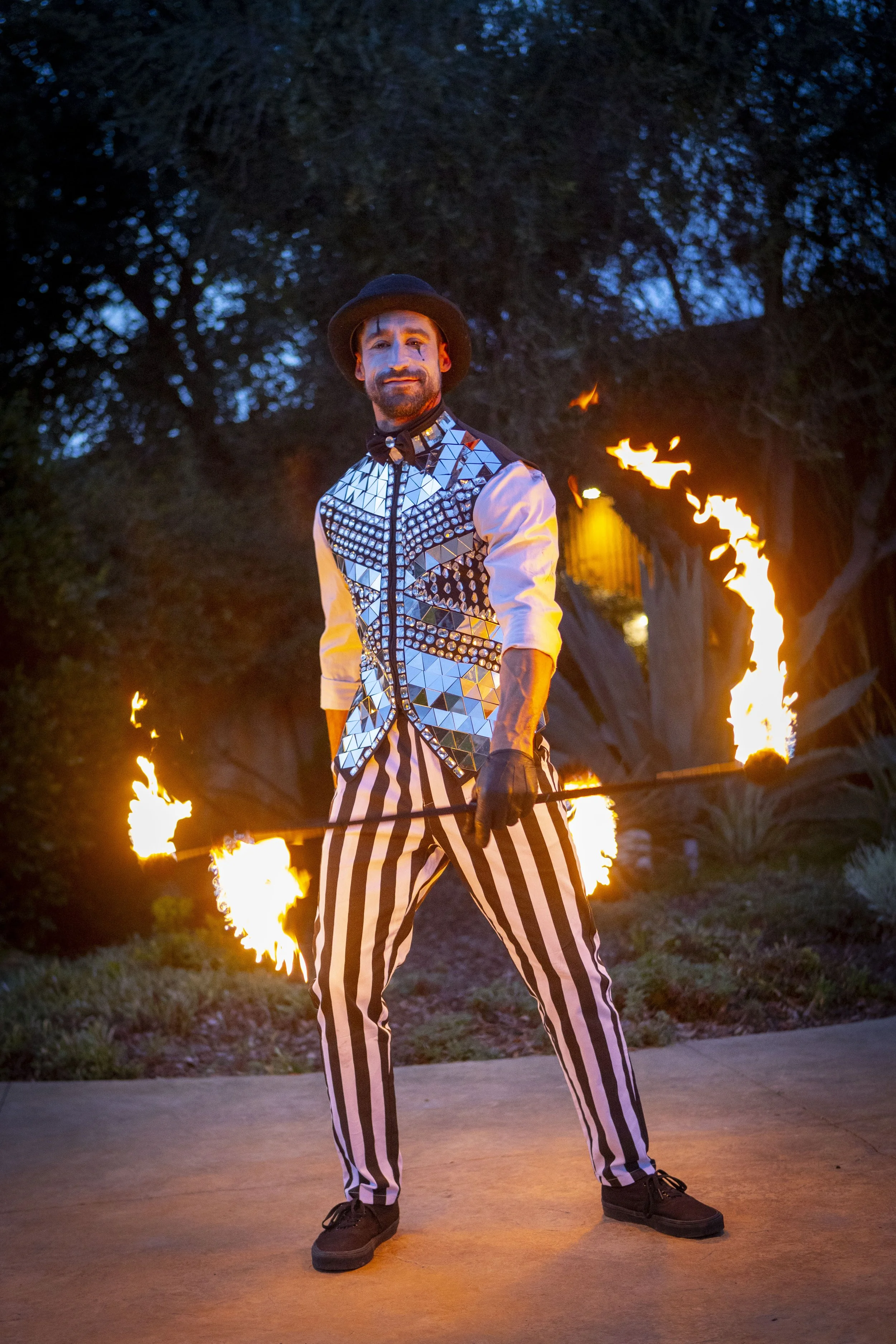 A male fire performer standing outdoors at night, holding lit fire torches, dressed in a reflective, geometric-patterned vest, striped pants, and a hat.