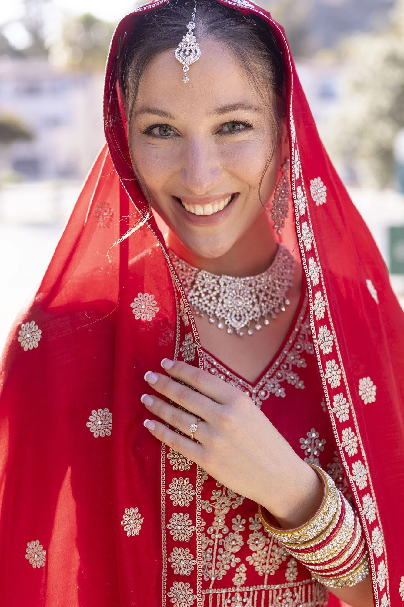 A woman smiling in traditional Indian bridal attire, wearing a red saree with silver embroidery, embellished jewelry including a necklace, earrings, bangle, and a ring.
