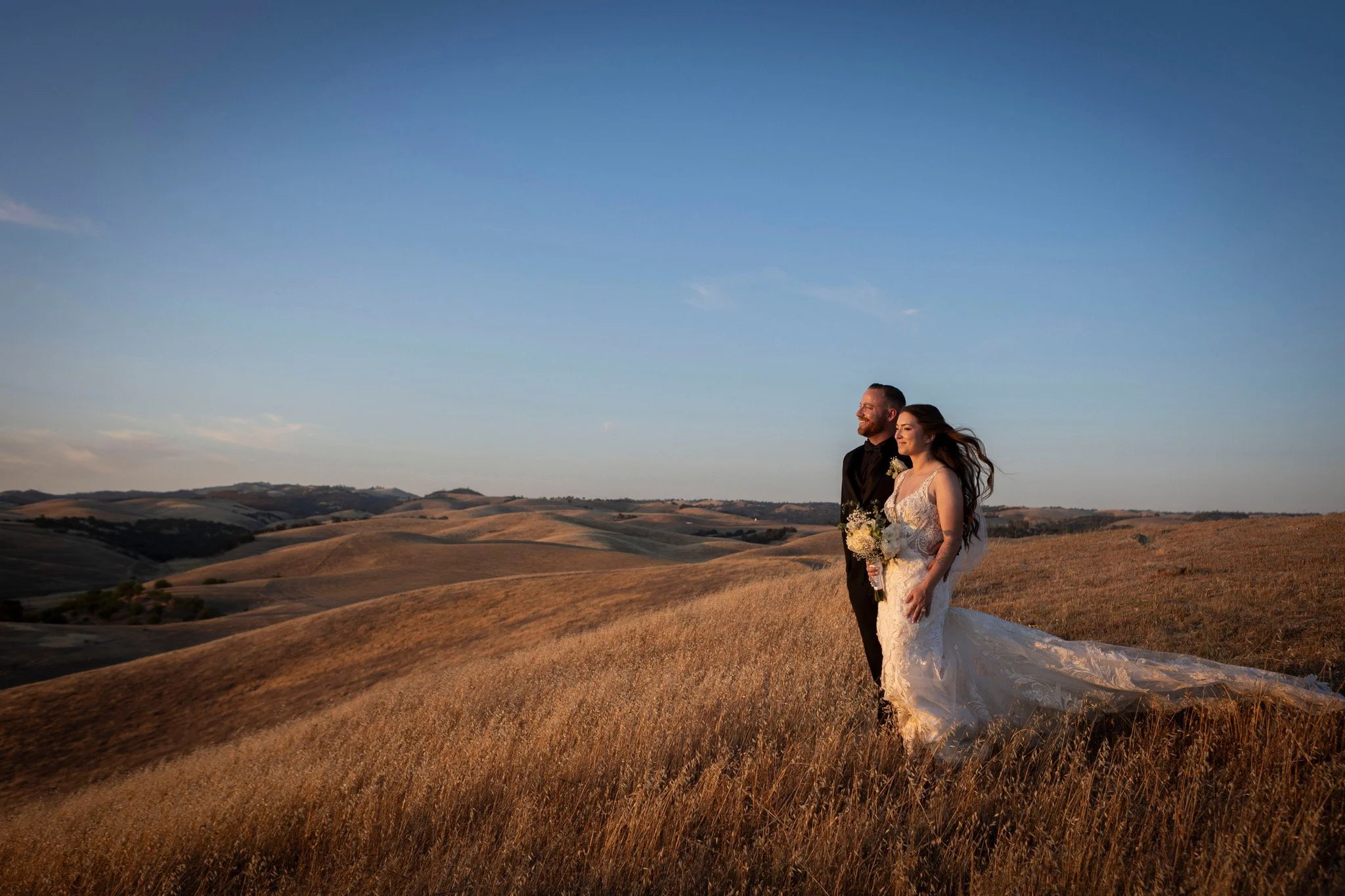 A bride and groom standing together in a field of tall, golden grass, with rolling hills in the background and a clear blue sky overhead, during sunset.