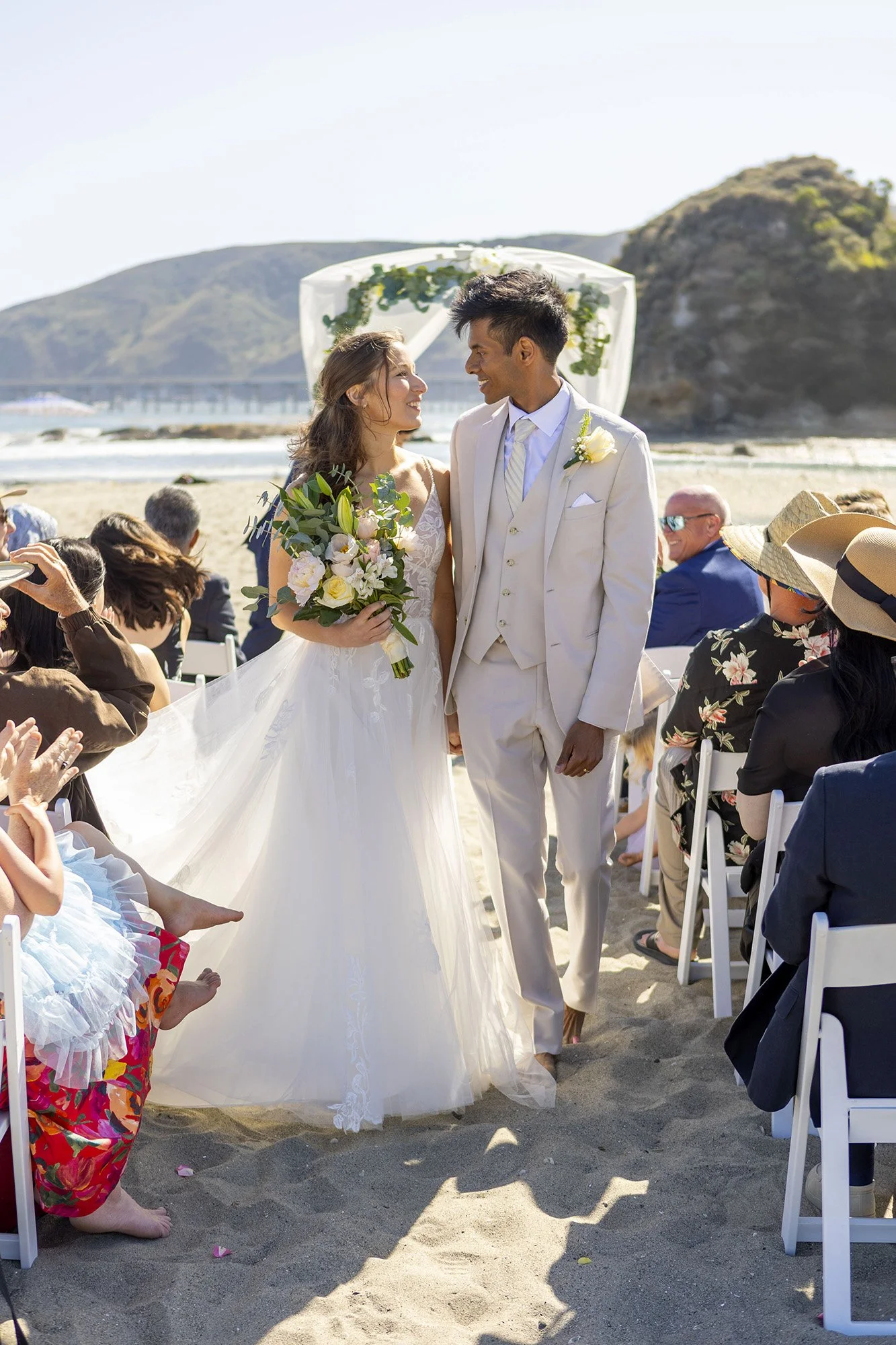 A bride and groom exchange vows at an outdoor beach wedding, with guests seated on chairs and scenic hills in the background.