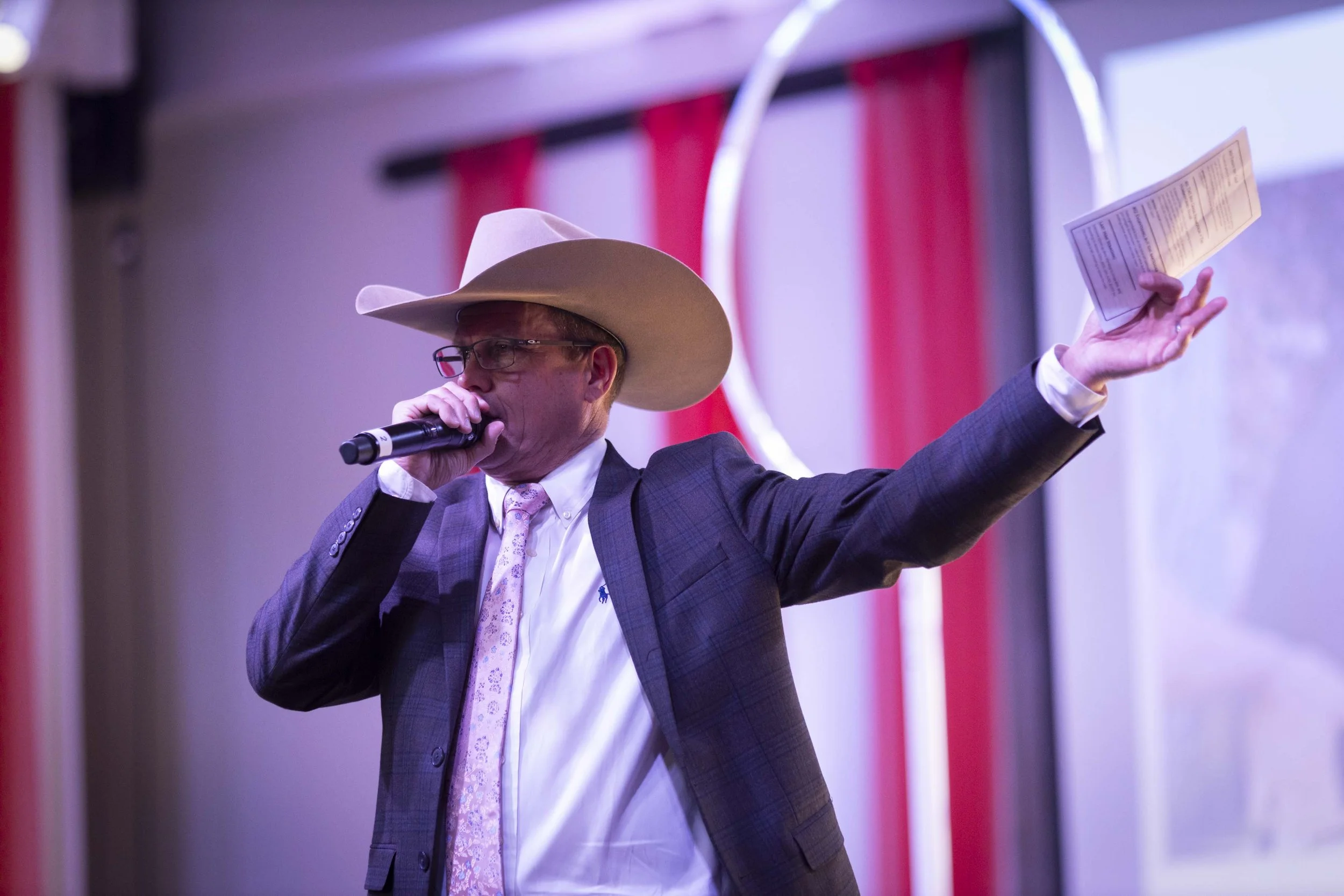 A man wearing a cowboy hat, glasses, a suit, and a tie, holding a microphone and a piece of paper, speaking or singing at an event.