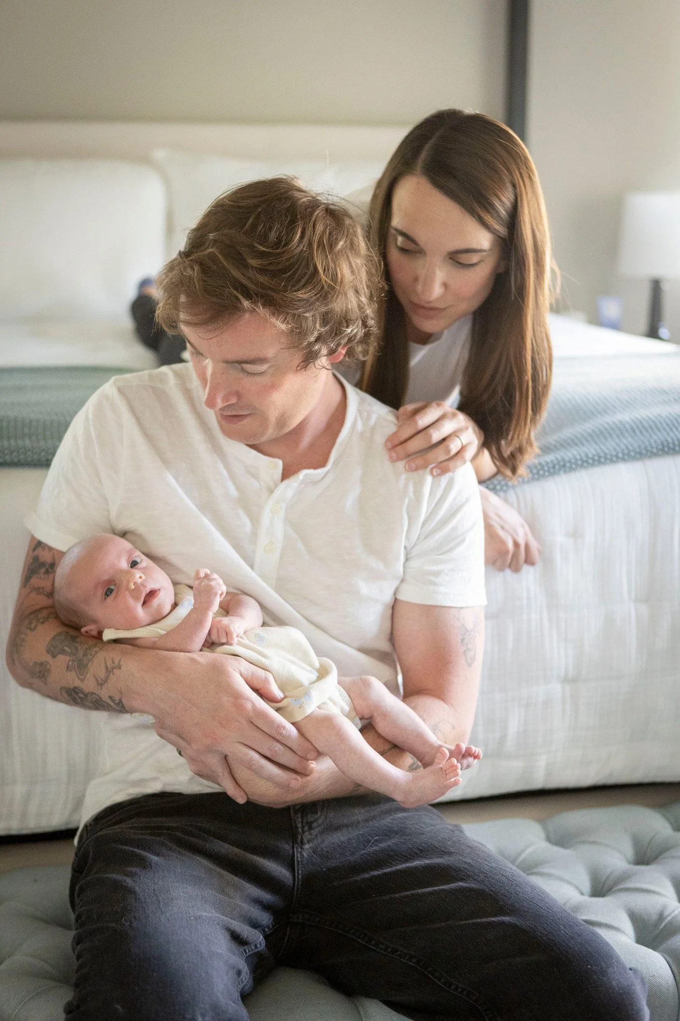 A man with tattoos holding a baby while a woman stands behind him, all in a bedroom.