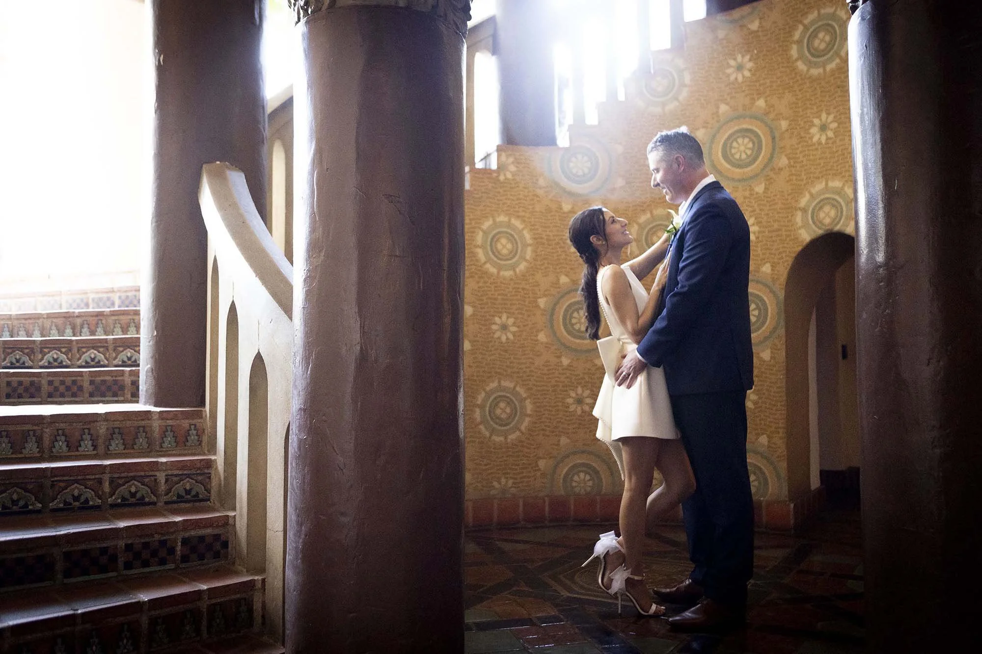 A bride and groom celebrating their wedding in an indoor setting with warm lighting, decorative wall, and staircase in the background.
