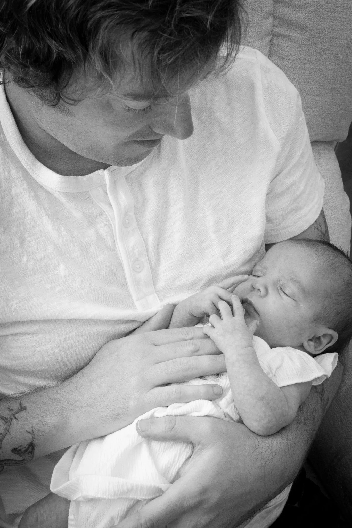 A man with dark, curly hair and a white shirt holding a sleeping baby in his arms, with a focus on their faces.