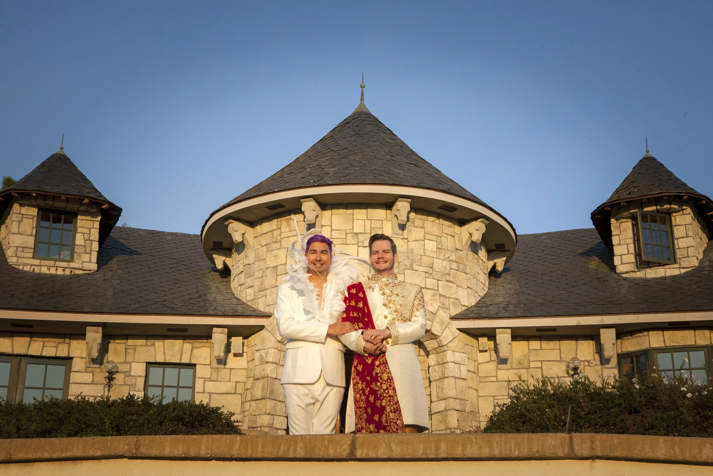 Two men in ornate, traditional Indian wedding attire standing in front of a castle-like stone building during sunset.