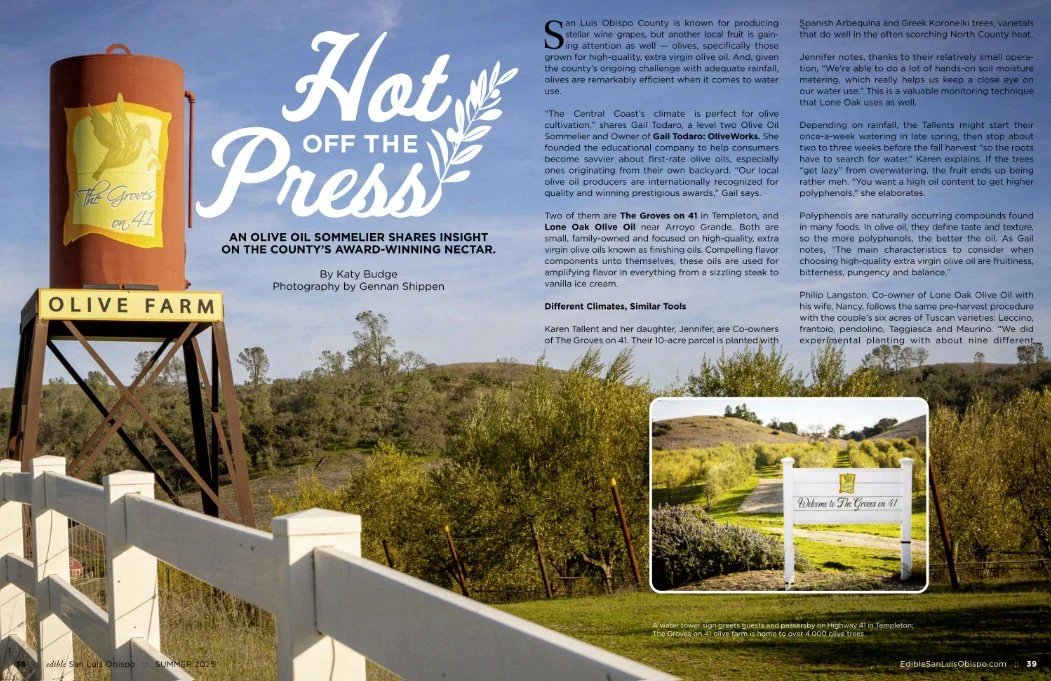 Sign for Olive Farm next to a white fence and a water tower, with a scenic view of olive trees and rolling hills in the background.