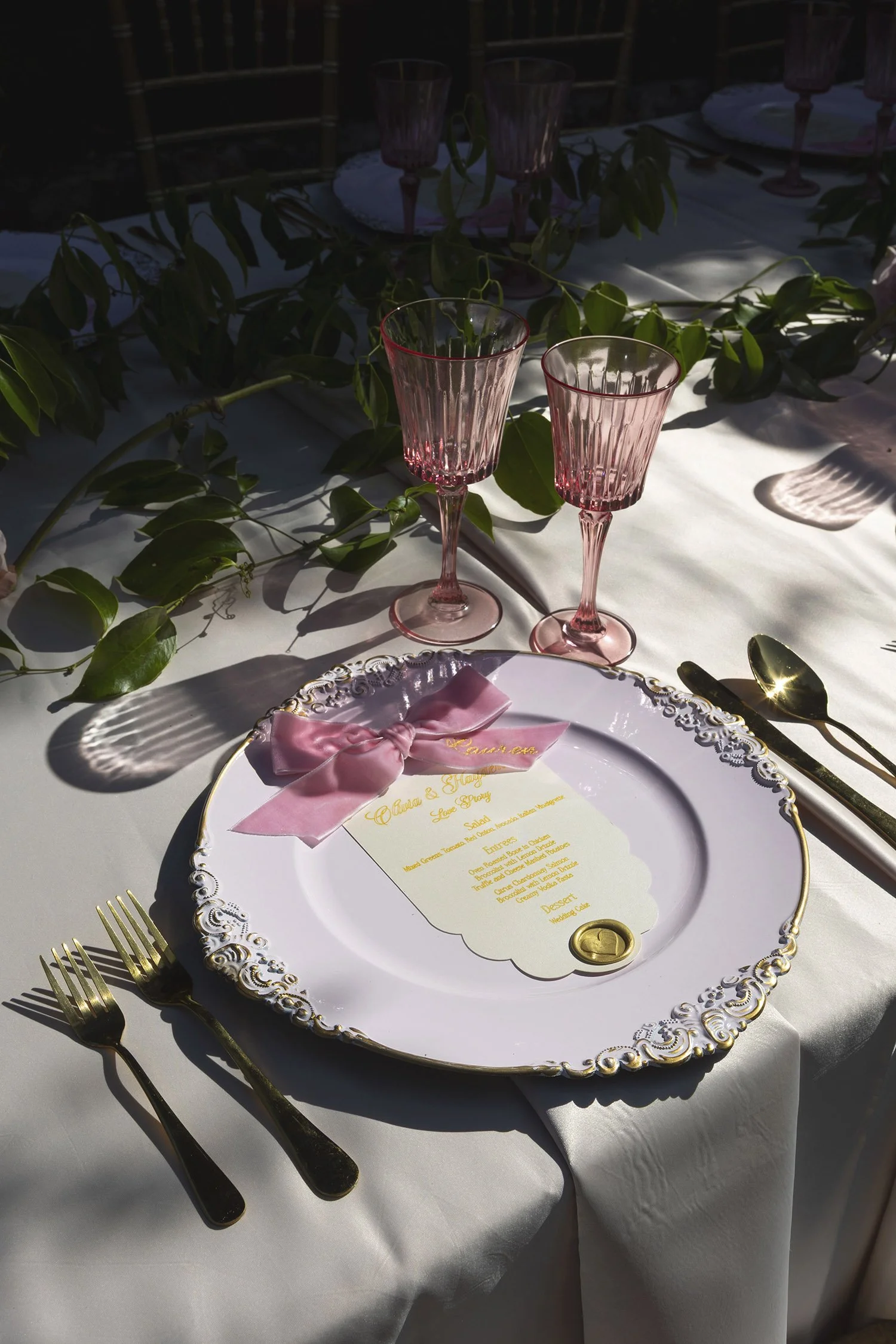 A beautifully set dining table with pink glassware, gold cutlery, a white ornate charger plate, a pink bow, and a printed menu in gold lettering on a white tablecloth, with leafy greenery decor.