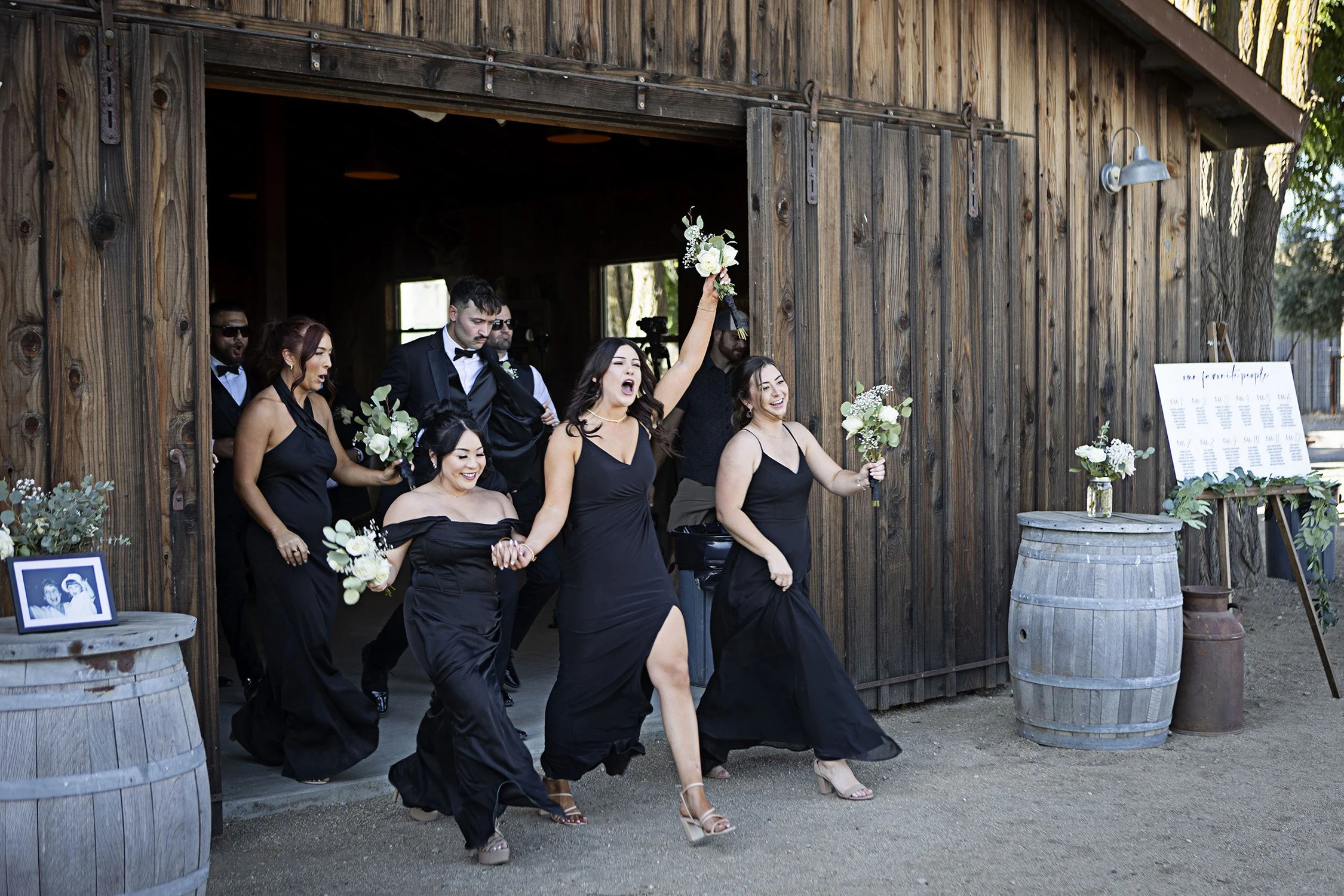 Wedding party in black dresses and tuxedos exiting a rustic barn, celebrating with smiles and raised bouquets.
