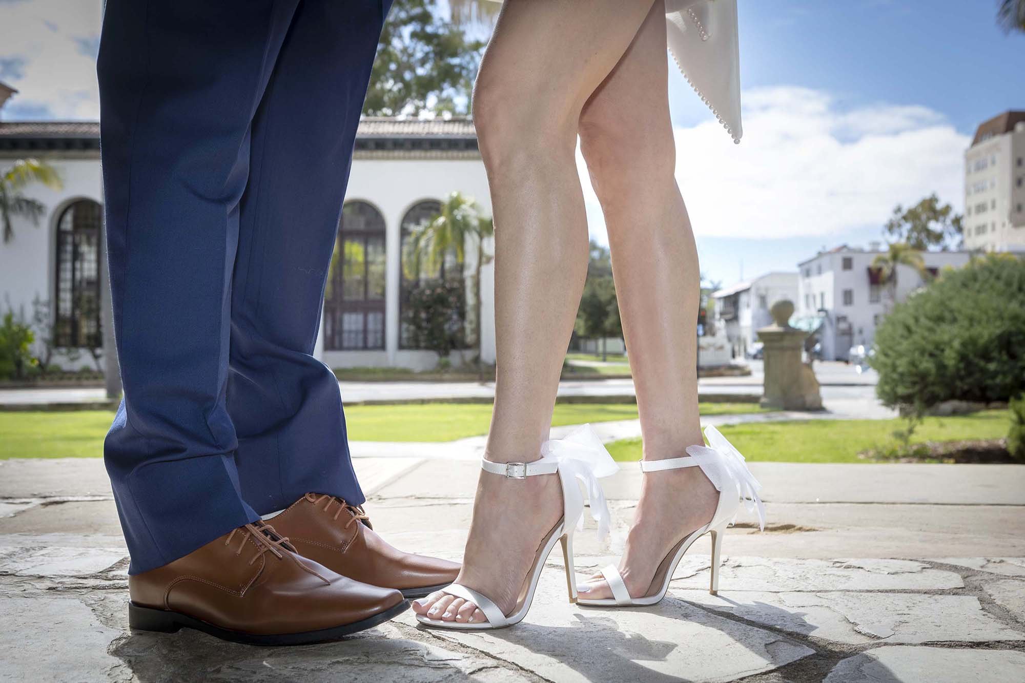 Close-up of a bride and groom's feet standing outdoors, bride in white high heels with bows, groom in brown dress shoes, cityscape in the background.