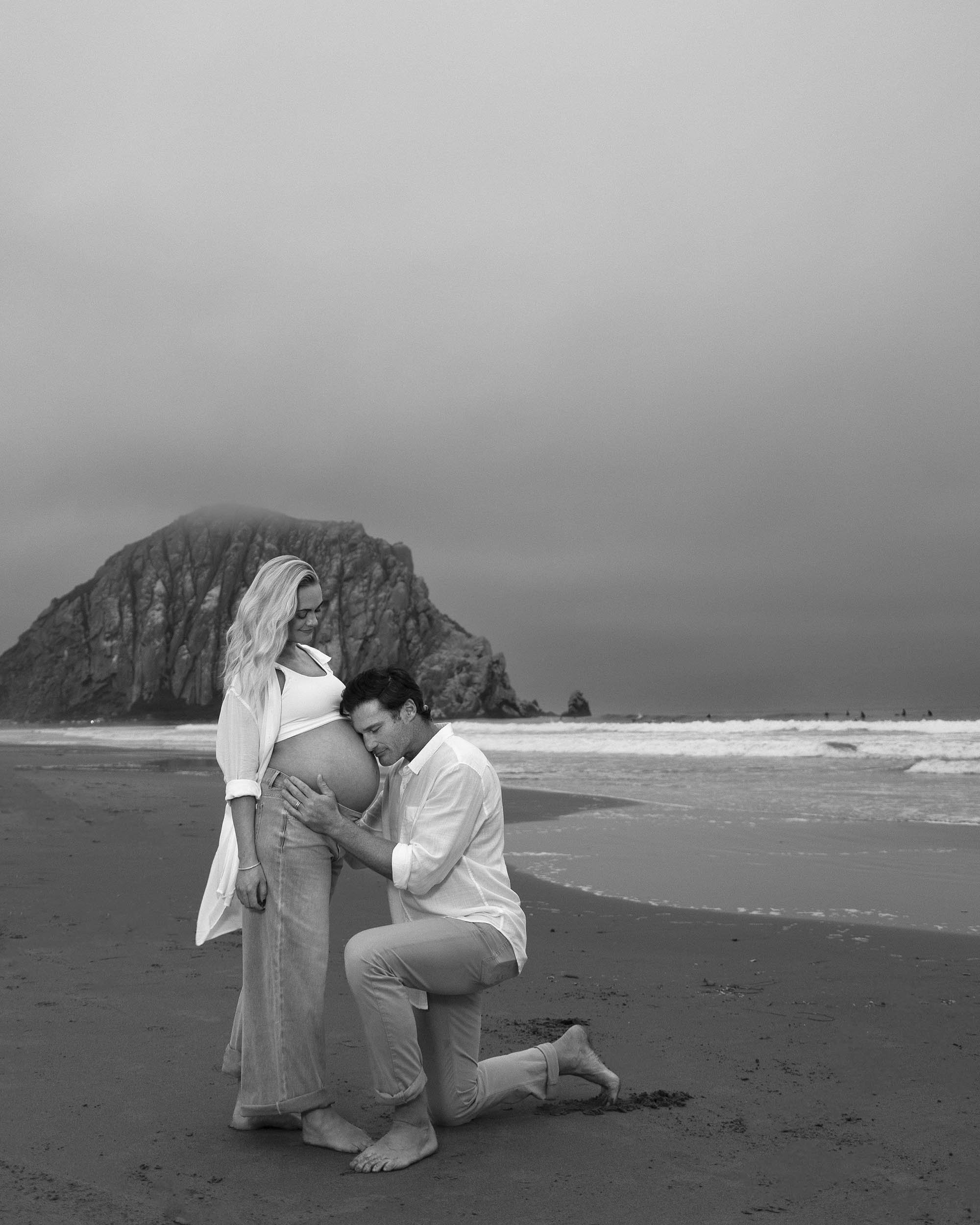 A couple is on a beach during sunset, with the woman kneeling and the man counting her belly, possibly during a pregnancy. The woman has long, wavy hair and is wearing a loose shirt, while the man has short hair and is wearing a button-up shirt. The 