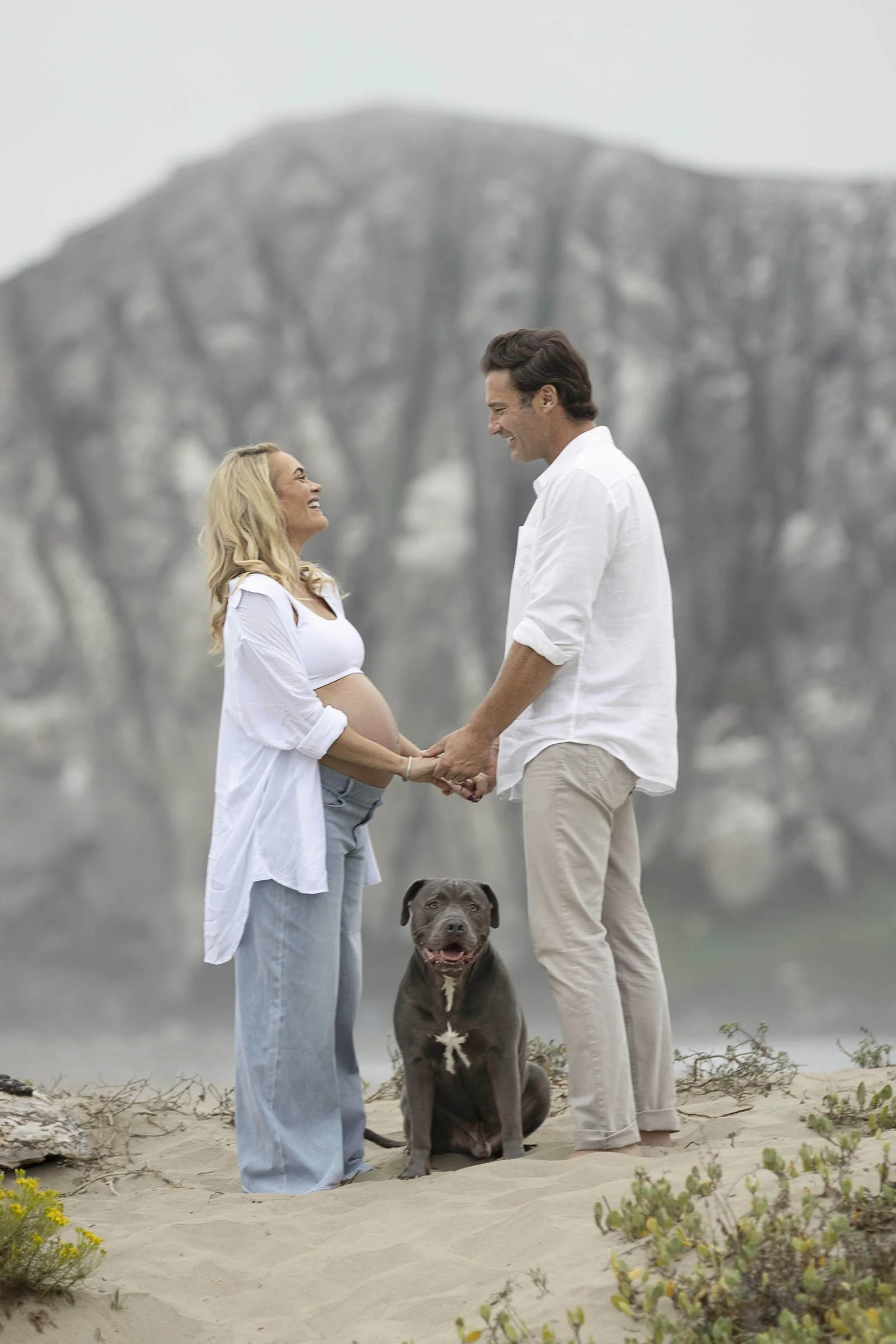 A pregnant woman and a man holding hands and smiling at each other on a beach with a dog sitting between them. The woman is wearing a white shirt and light blue jeans, the man is in a white shirt and beige pants. They are outdoors with a mountain in 