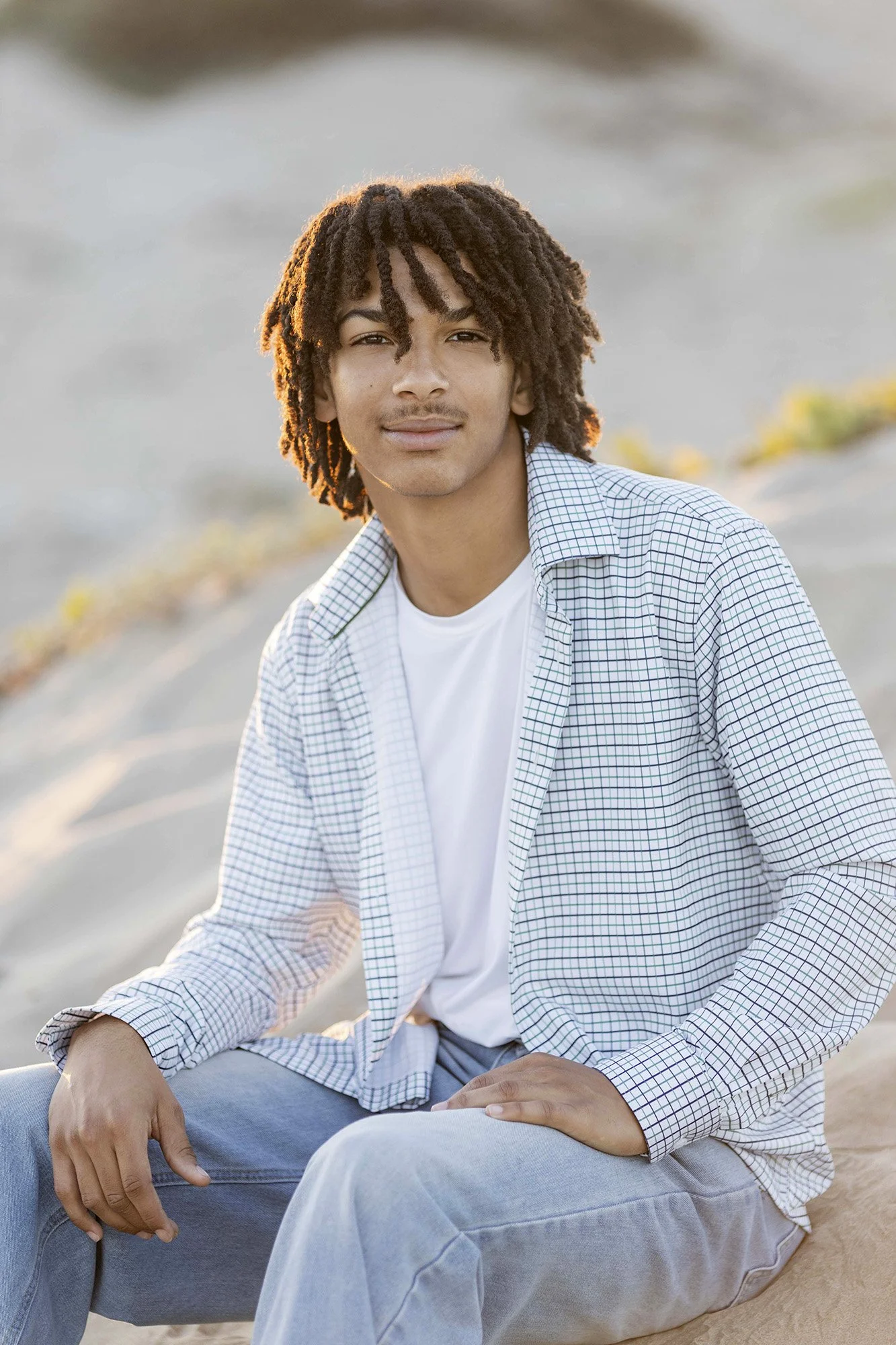 A young man with curly hair sitting outdoors near water, wearing a white T-shirt and a checked shirt.