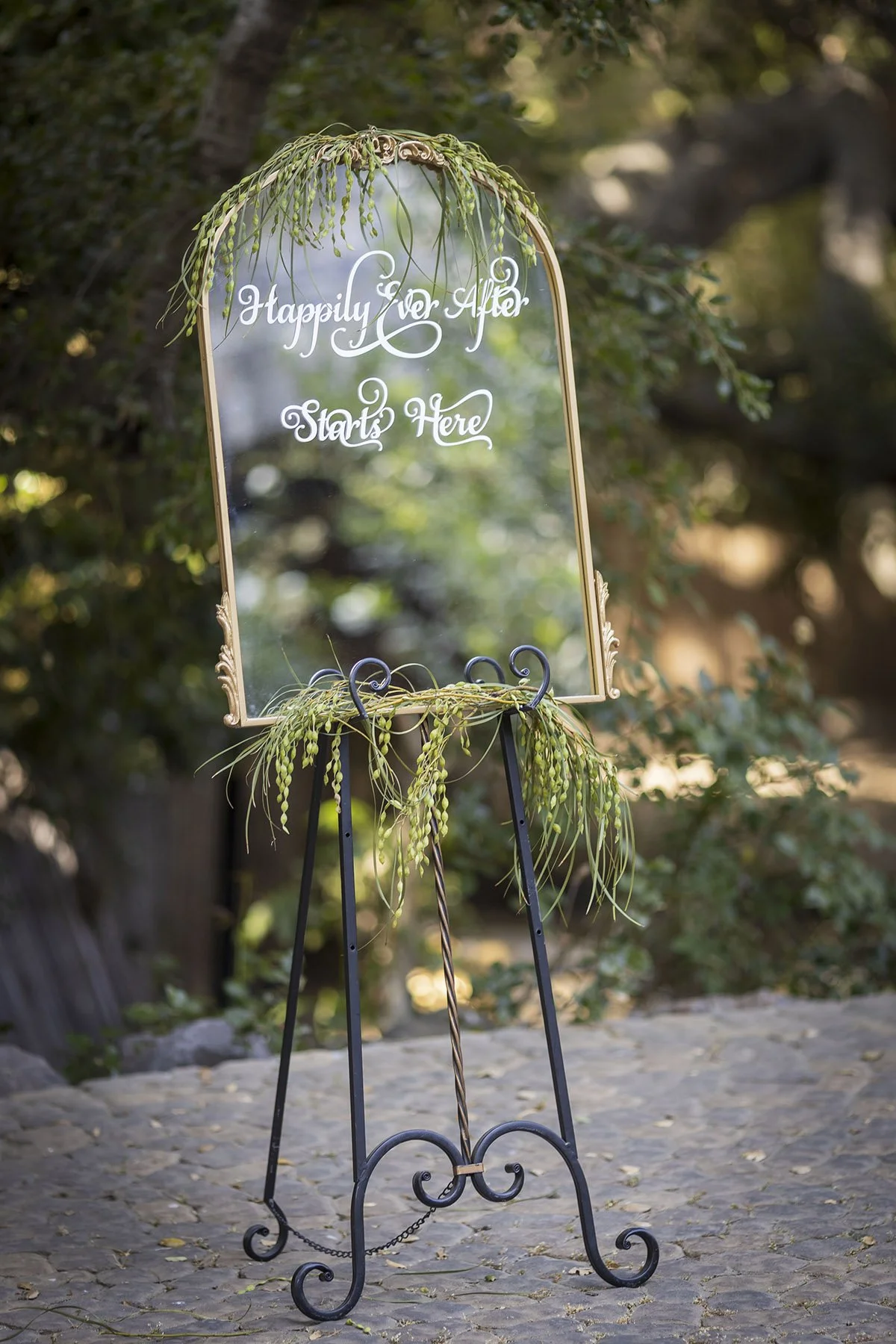 A decorative mirror with a gold frame and script writing that says "Happily Ever After Starts Here" on it. The mirror is adorned with greenery on top and bottom, and is placed on a black metal stand outdoors on a stone surface surrounded by trees and