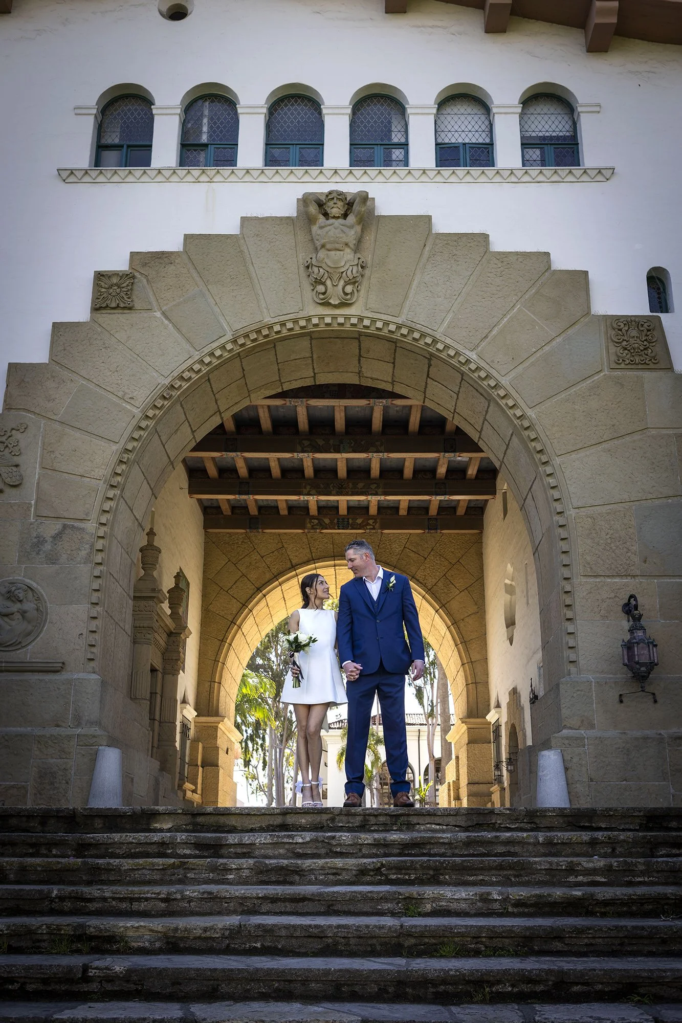 A bride and groom holding hands and walking down stone steps in front of an archway, with a historic building featuring ornate stonework and narrow windows in the background.