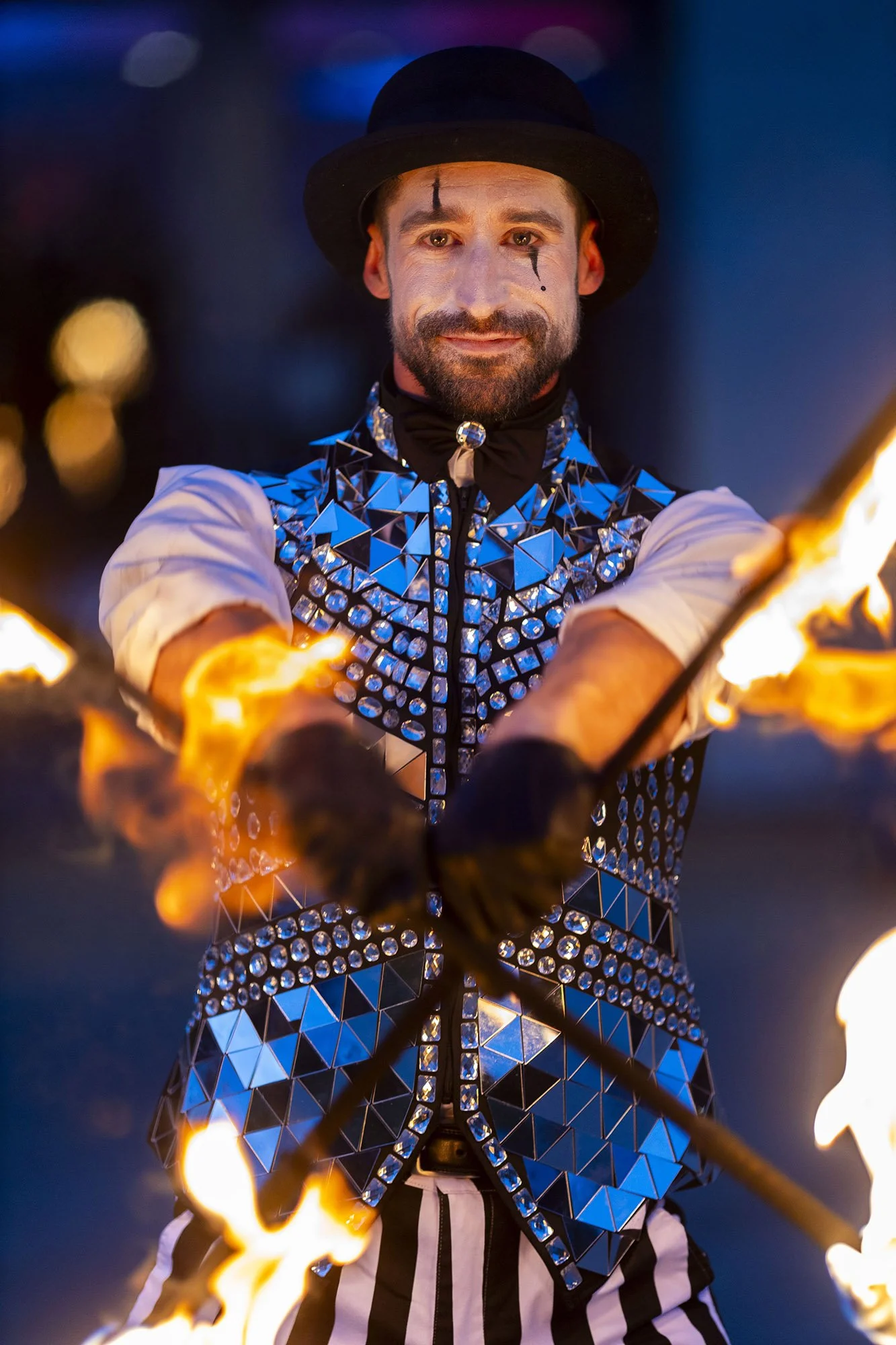 A male fire performer in a mirrored costume, black bowler hat, and striped pants, holding a flaming torch, smiling at the camera during a night event.