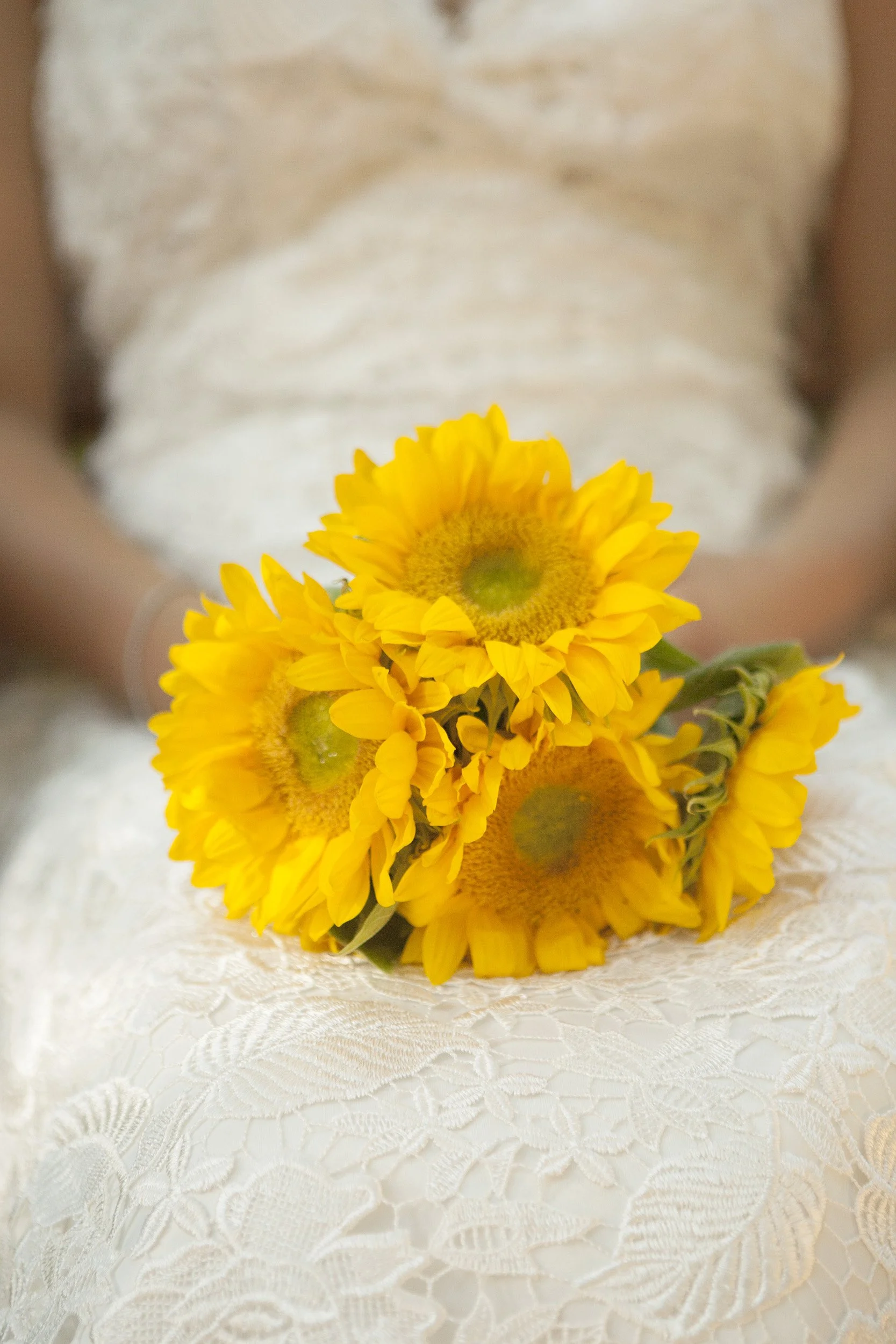 A person in a white lace dress holding a bouquet of bright yellow sunflowers.