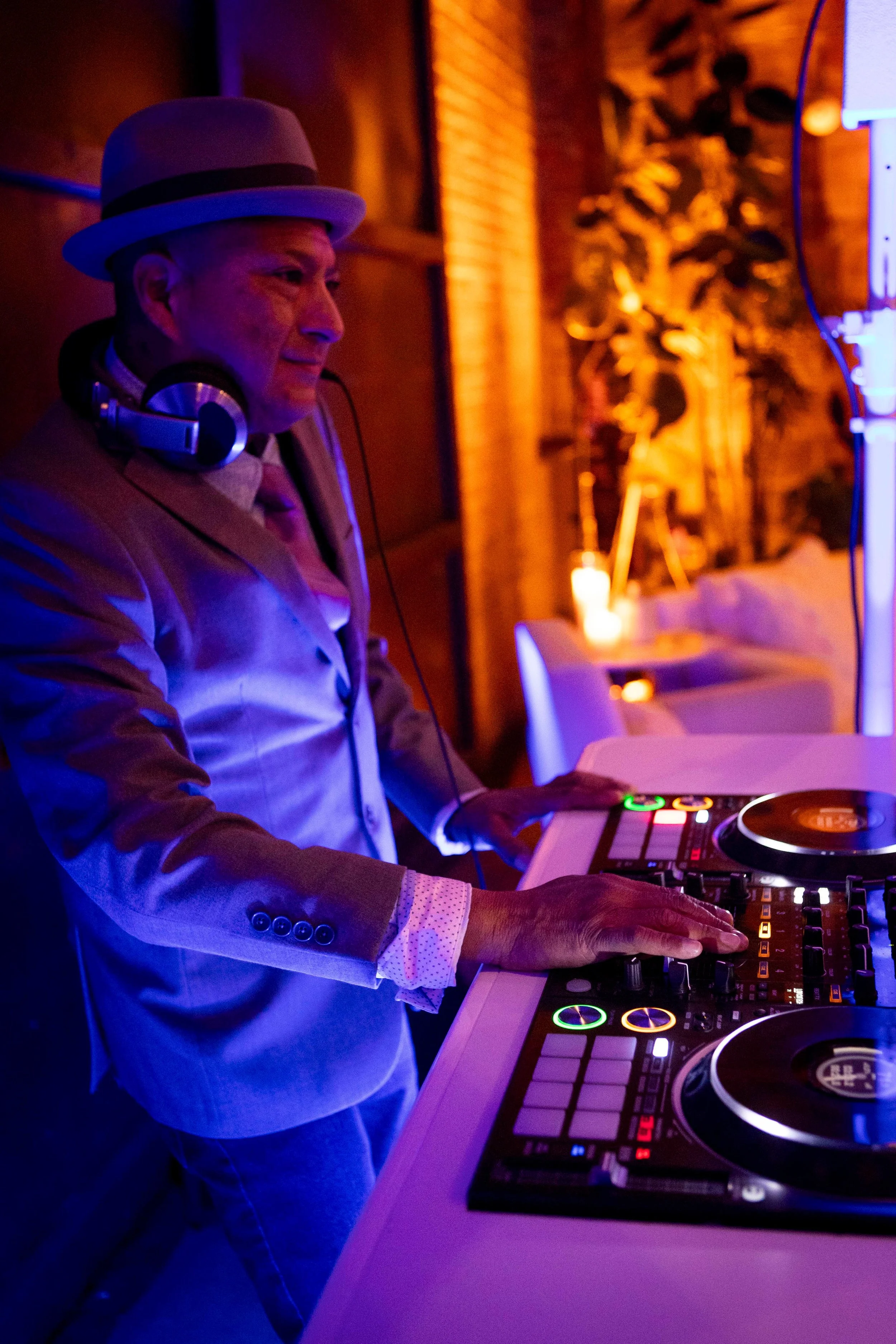 A man wearing a light-colored suit and a fedora hat DJing at an event with colorful lighting, using a DJ controller and wearing headphones.