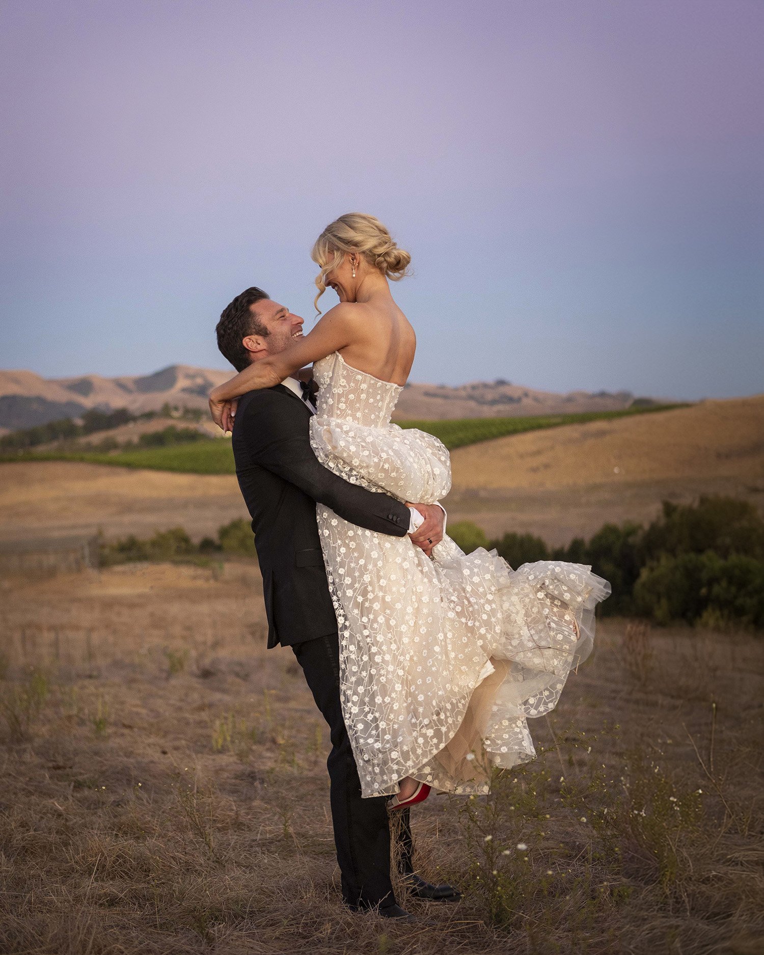 A groom lifting a bride in a field during sunset