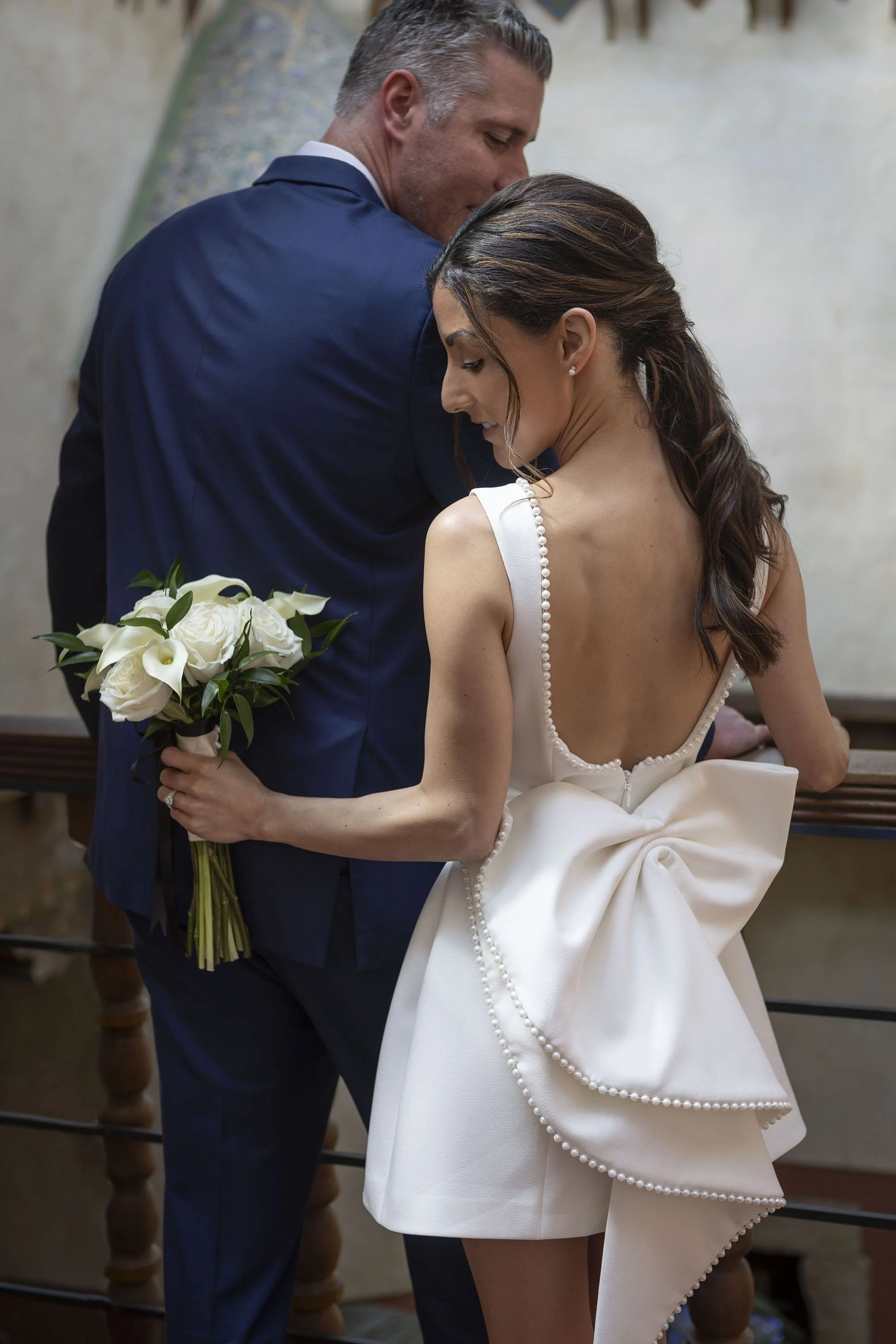 A man in a blue suit and a woman in a white dress sharing a close, intimate moment at a wedding, with the woman holding a bouquet of white roses and calla lilies, both with their eyes closed.