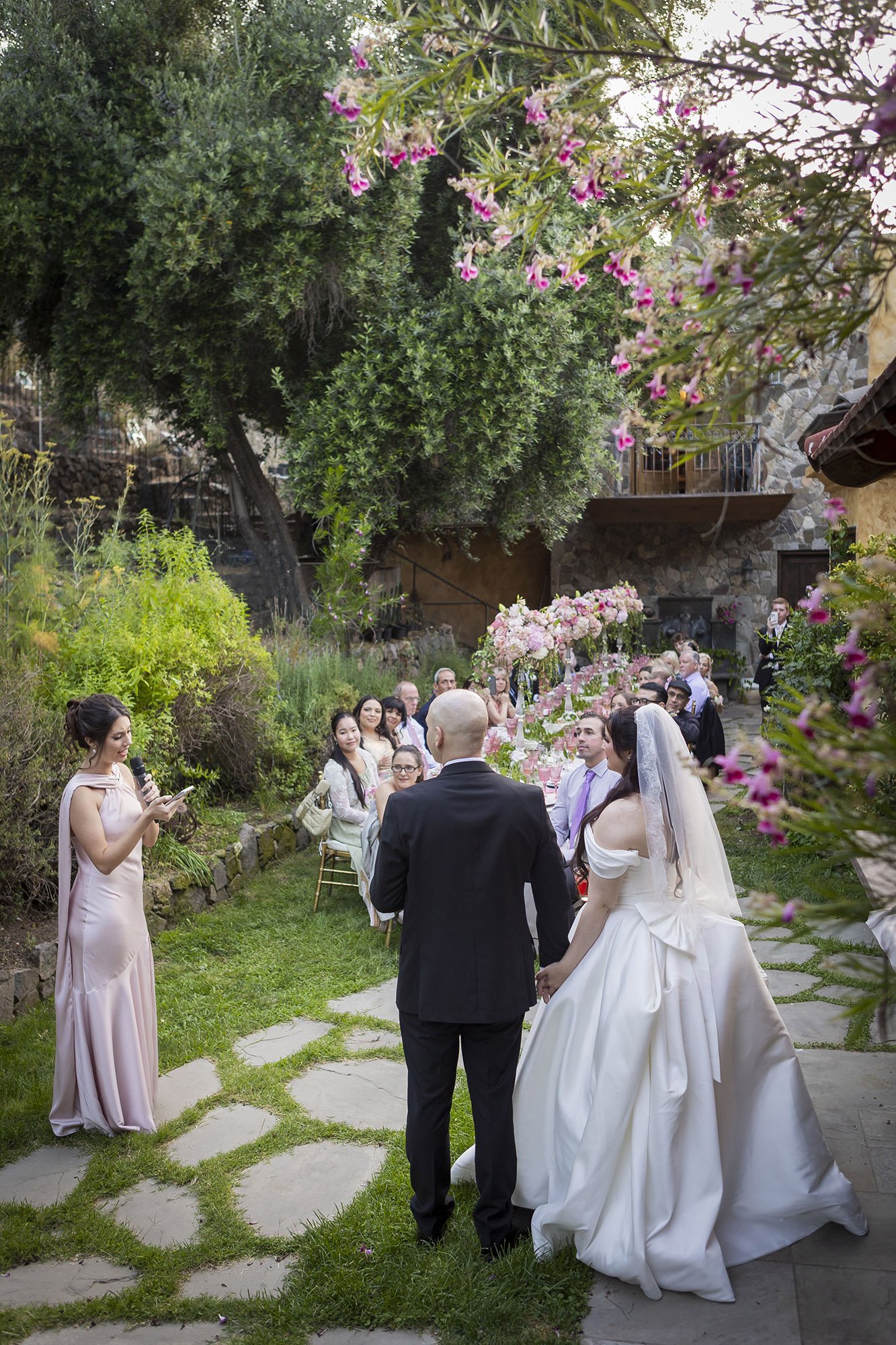 A wedding ceremony taking place outdoors in a lush garden, with a bride and groom holding hands facing an officiant and seated guests, surrounded by greenery and pink flowers.