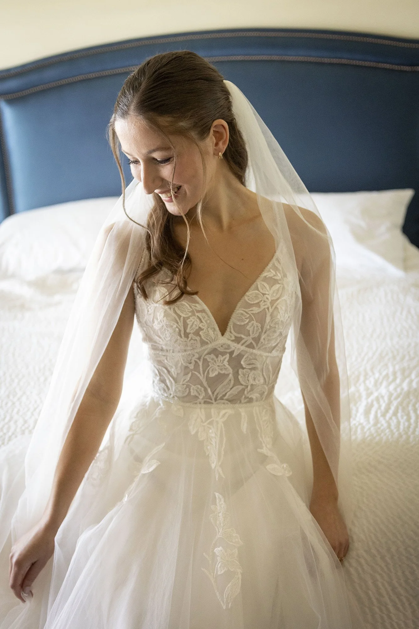 A bride in a wedding dress sitting on a bed, smiling and looking down.
