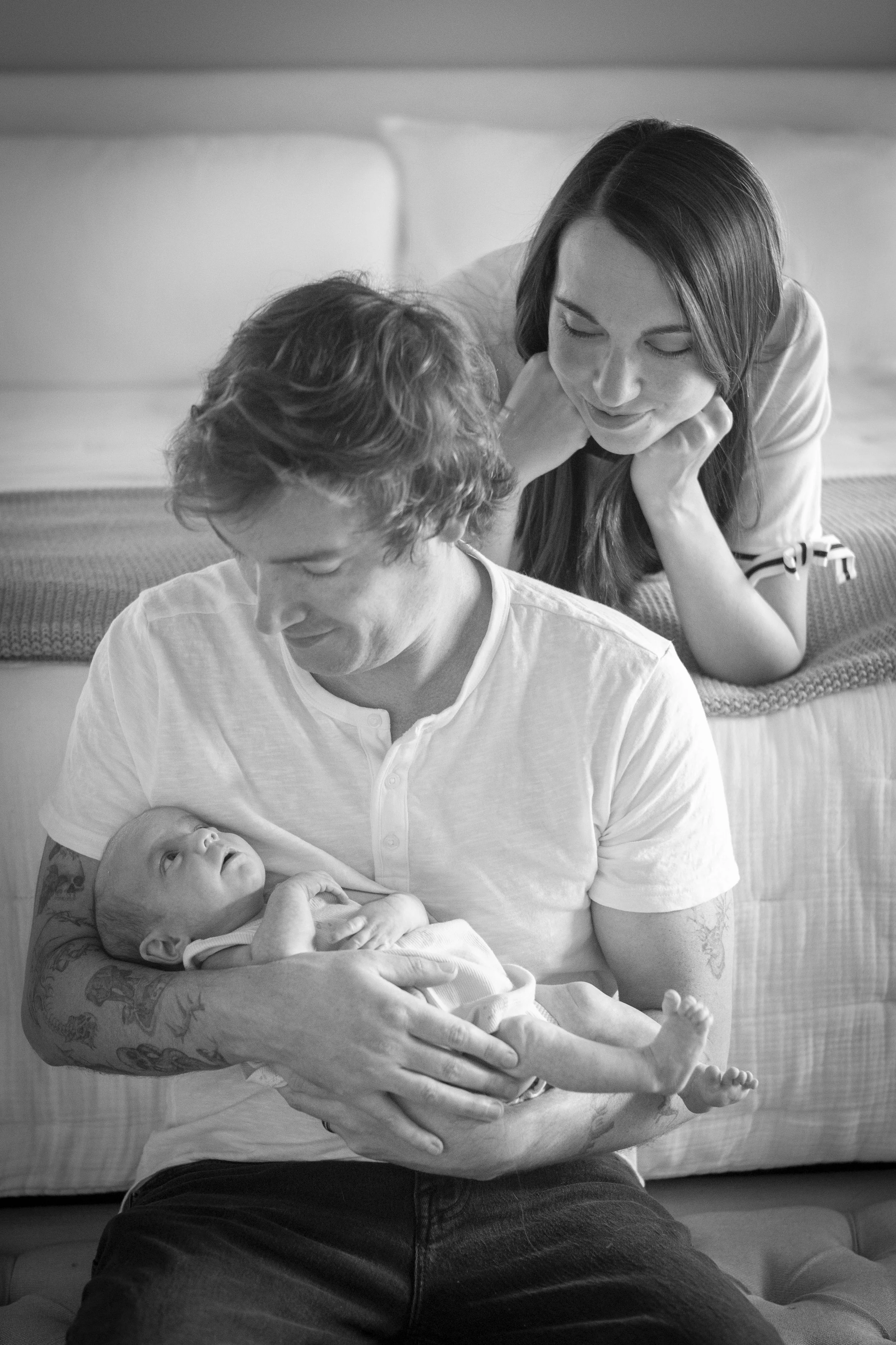 A woman is lying on her stomach on a bed, gently touching her face with her hand, and looking down at a man who is sitting and holding a baby in his arms. The woman appears to be smiling softly at the baby, creating a tender family scene.