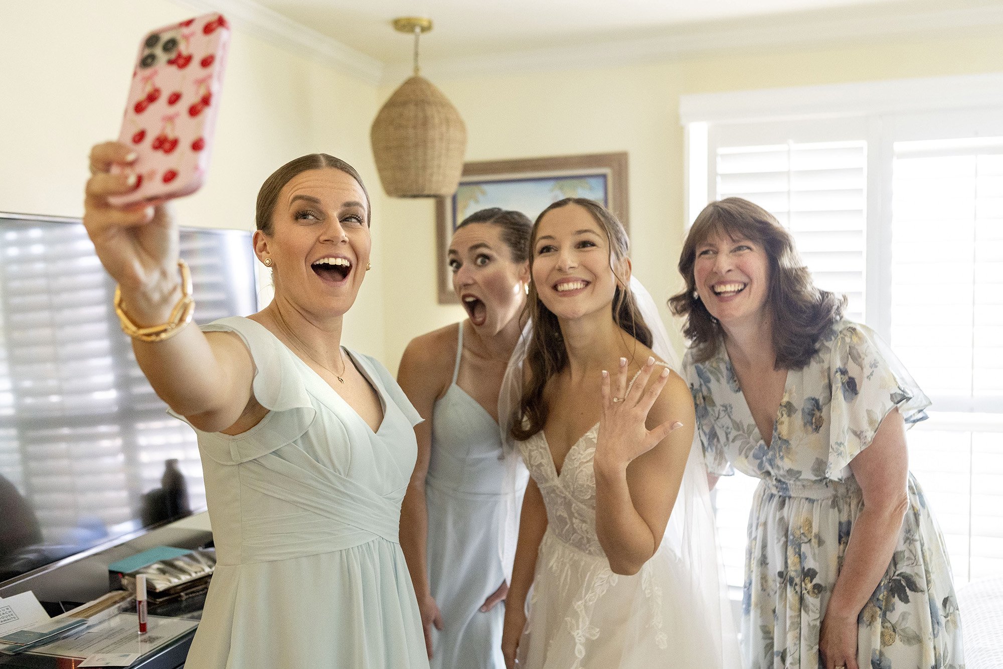 Group of four women taking a selfie, one bride wearing a white wedding dress, and others in pastel dresses, inside a bright room with window blinds and a painting on the wall.