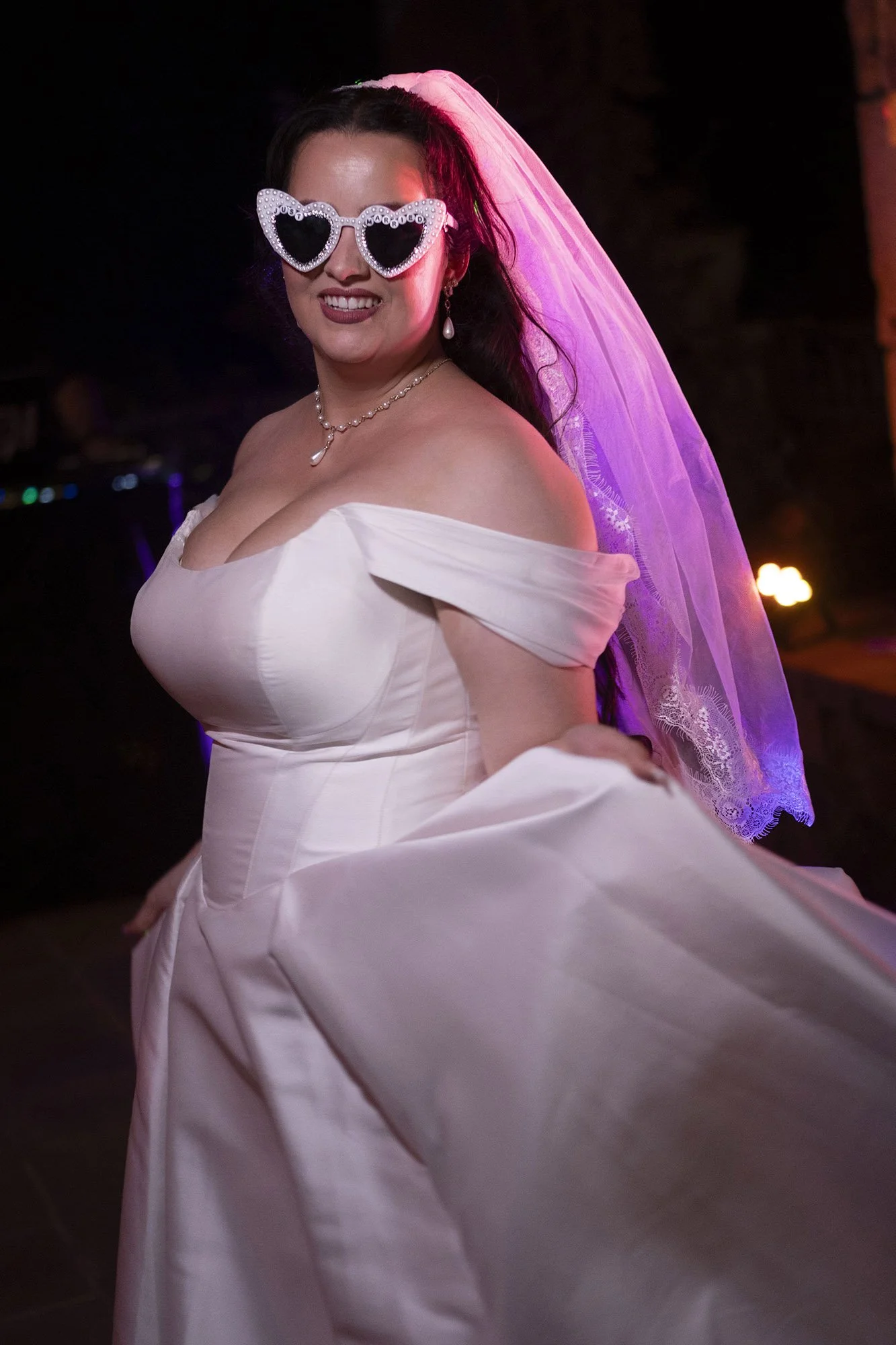 A woman in a white wedding dress, wearing heart-shaped sunglasses with pearl embellishments, a veil, and jewelry, smiling at the camera in a dimly lit setting.