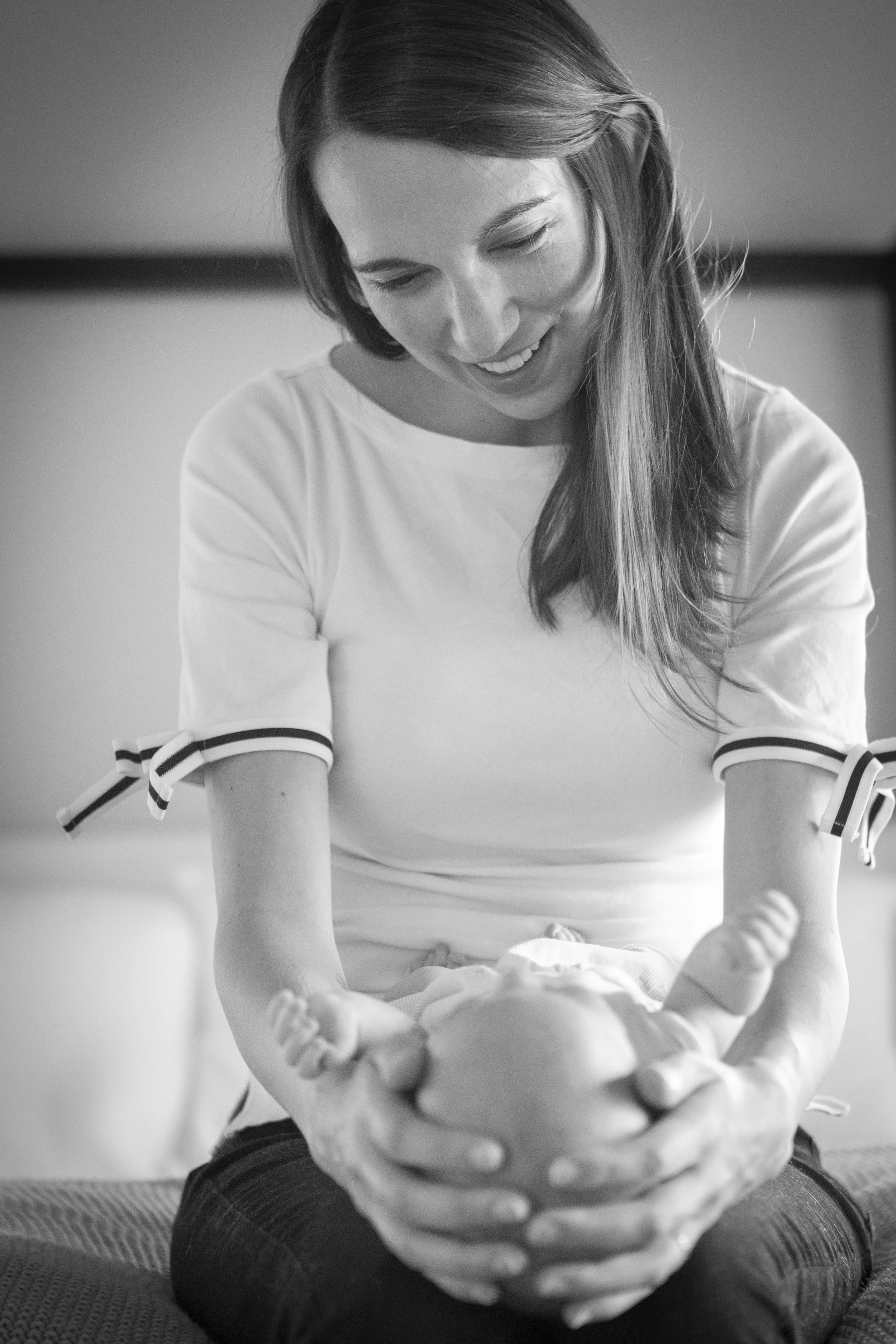 A woman with long hair smiling warmly while leaning forward, with her hands around a baby on her lap, in a cozy indoor setting. Black and white photo.