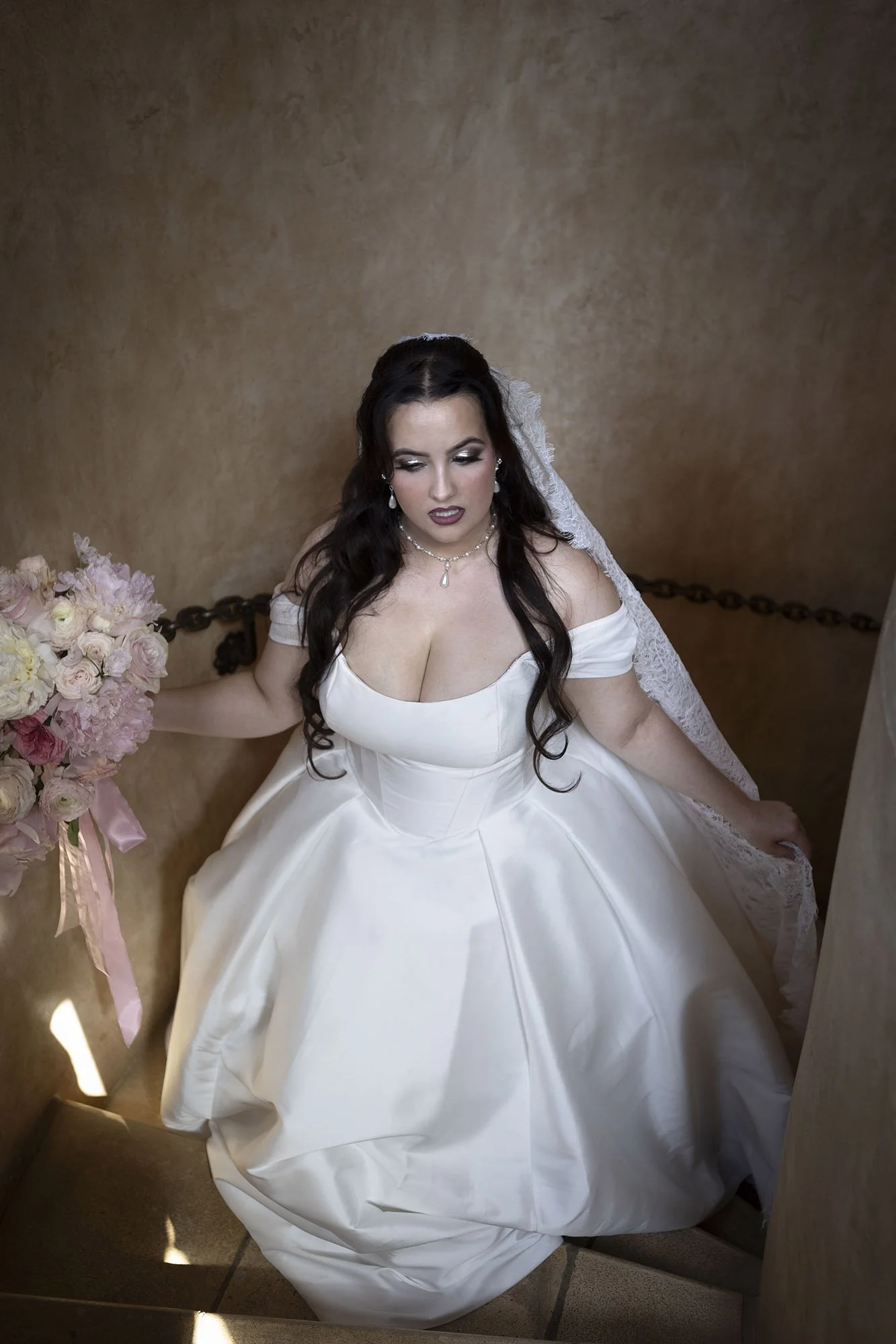 A bride in a white wedding dress sitting on a staircase, holding a bouquet of pink and white flowers, with lace on her veil, looking downward.