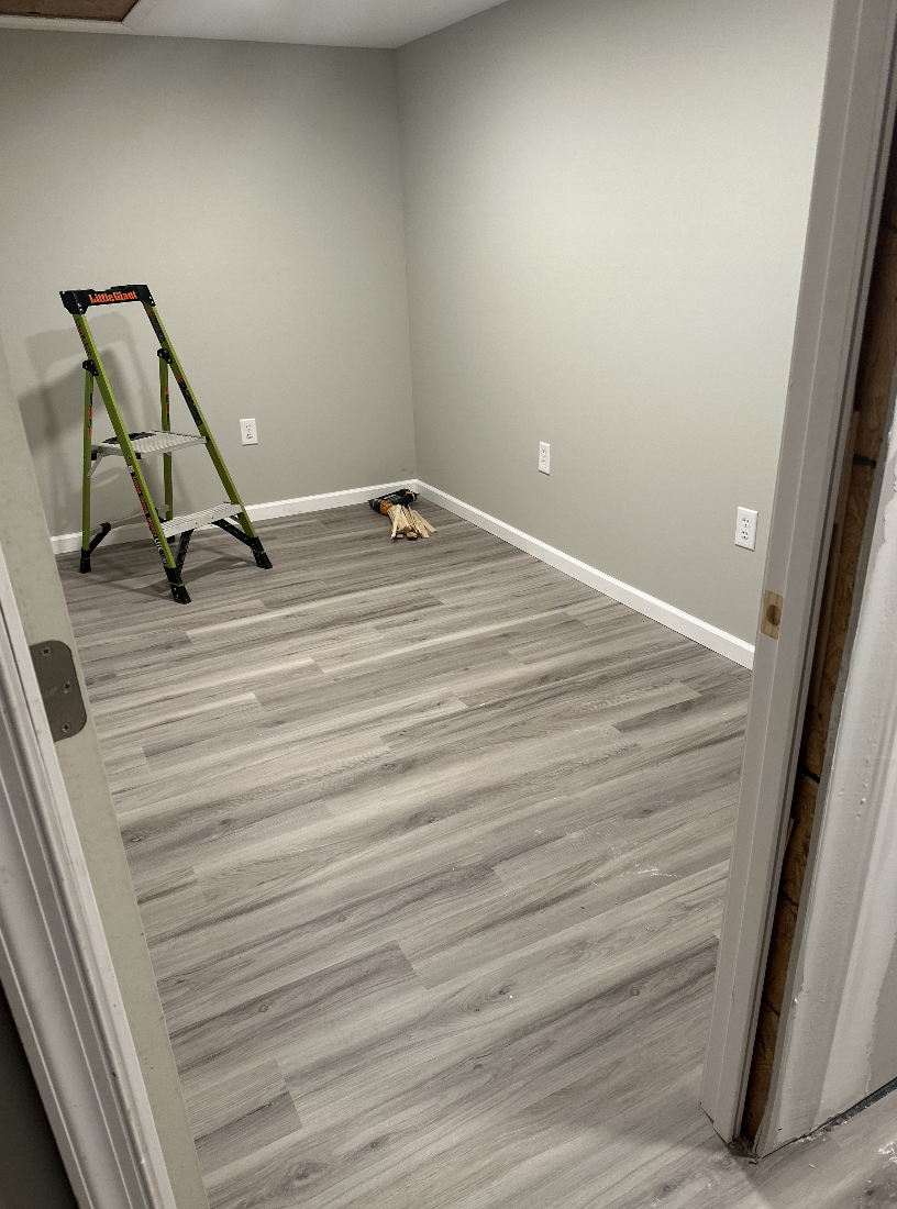 Empty room with light wood laminate flooring, gray walls, three electrical outlets, a green step ladder, and a pile of tools and wood pieces on the floor.