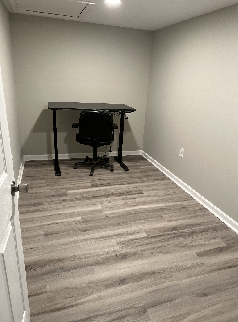 Small, empty room with light gray walls, wood laminate flooring, a closed door, a black desk, and a black office chair.