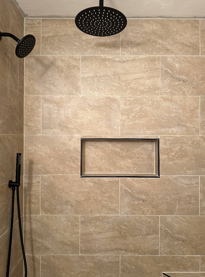 A walk-in shower with beige tiled walls, a round black rainfall showerhead at the ceiling, a black handheld shower on the left, and a built-in rectangular niche for toiletries.