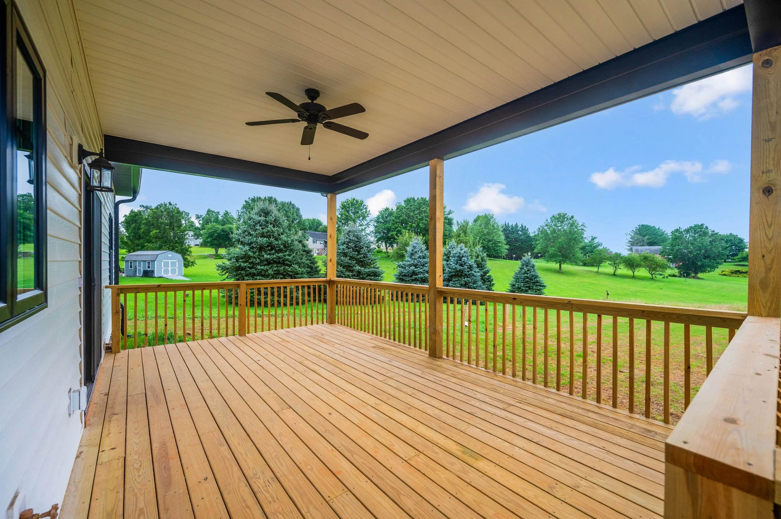 View of a wooden deck with railing overlooking a green yard with trees and a shed in the distance under a blue sky with white clouds.