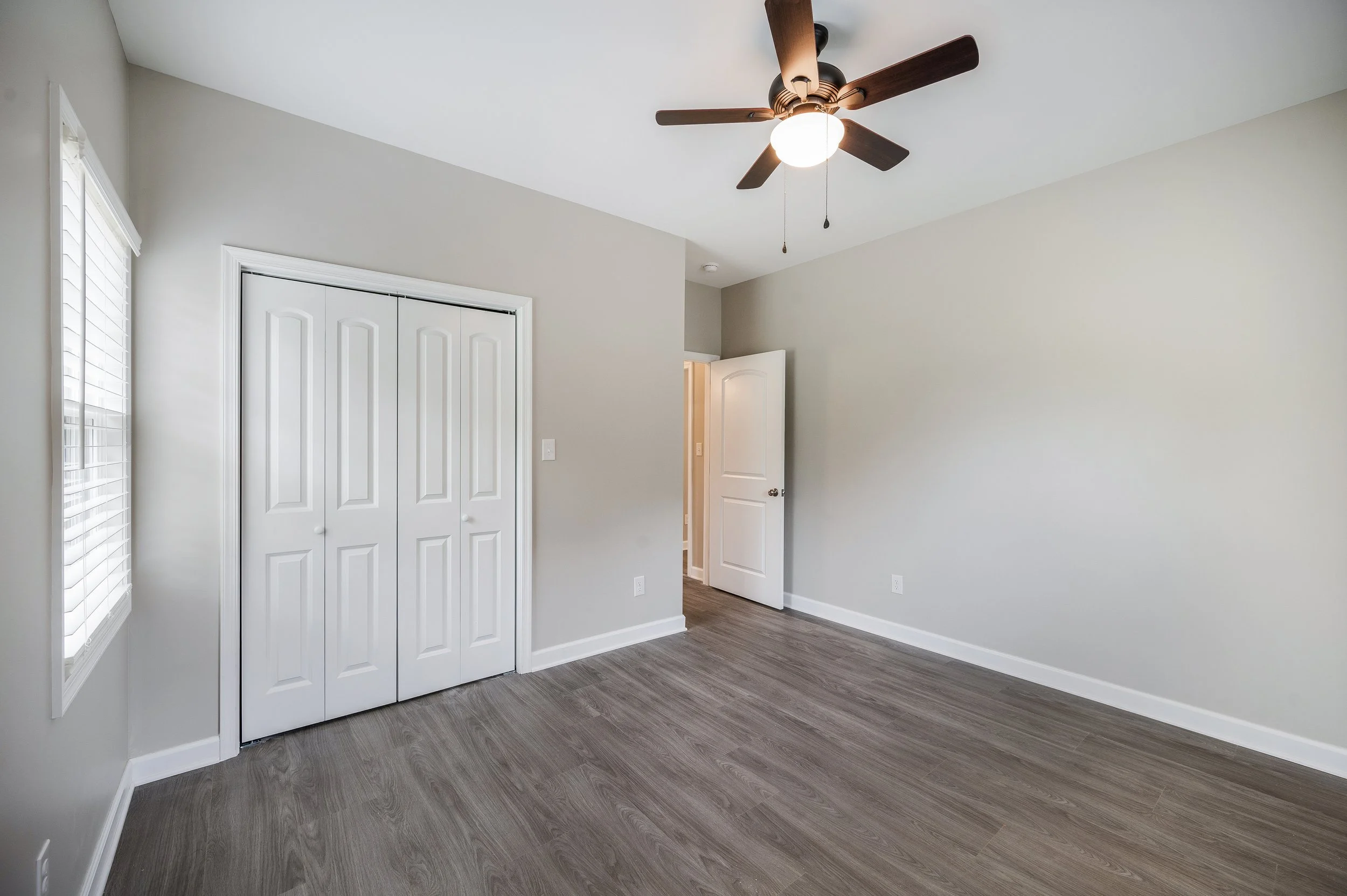 Empty bedroom featuring gray walls, wood-look flooring, a white closet with double doors, a ceiling fan with a light, and a window with white blinds.