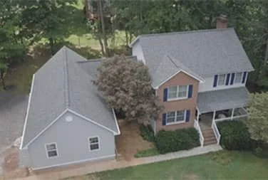 Aerial view of a residential house with a gray roof, brick and siding exterior, front porch, and front steps, surrounded by trees and a lawn.
