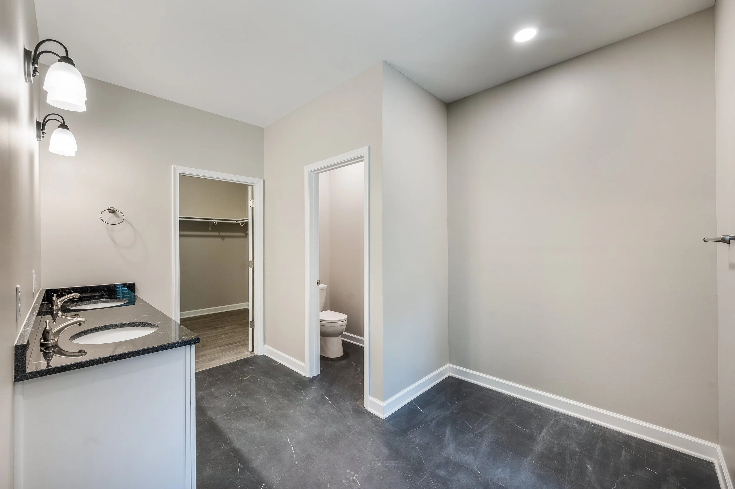 Empty bathroom with dual sinks, mirror lights, and a walk-in closet with a bar for hanging clothes.