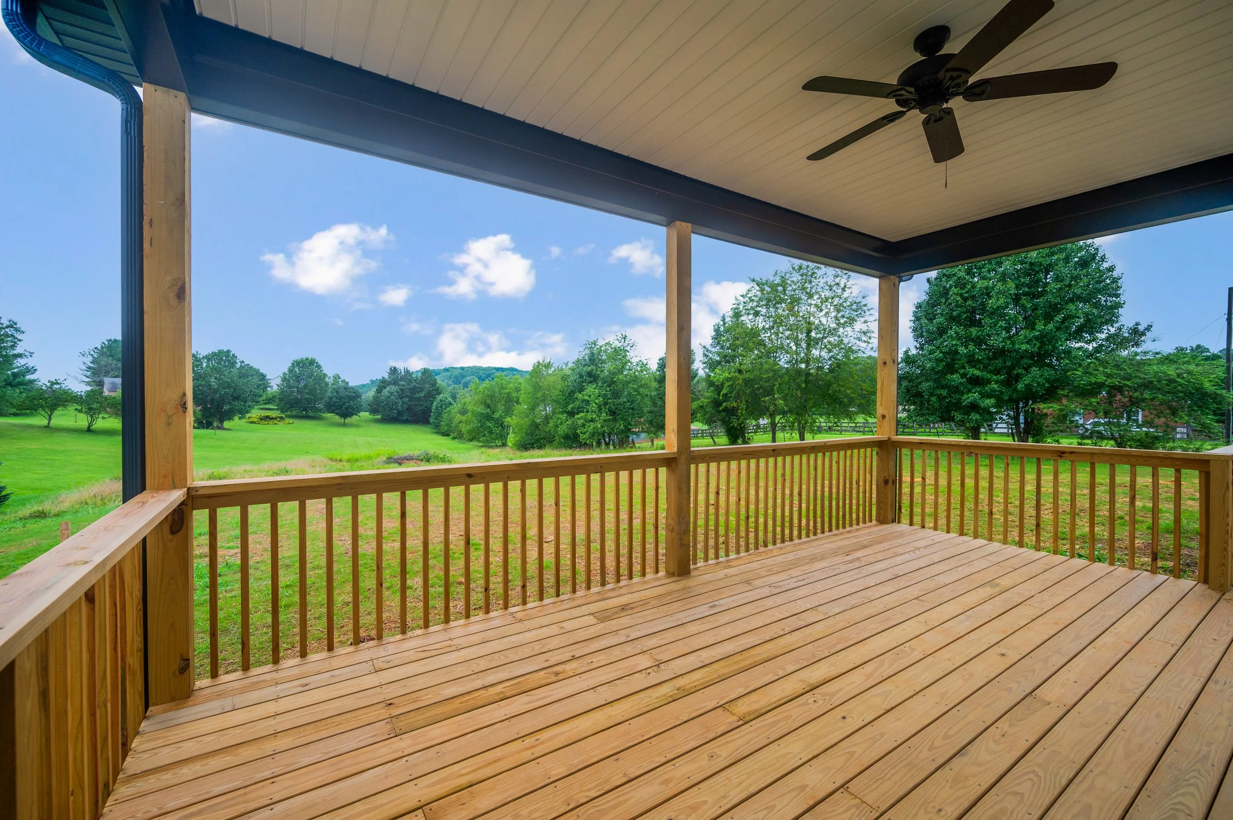View from a wooden porch with railing overlooking a green landscape with trees, grass, and a clear blue sky with a few clouds