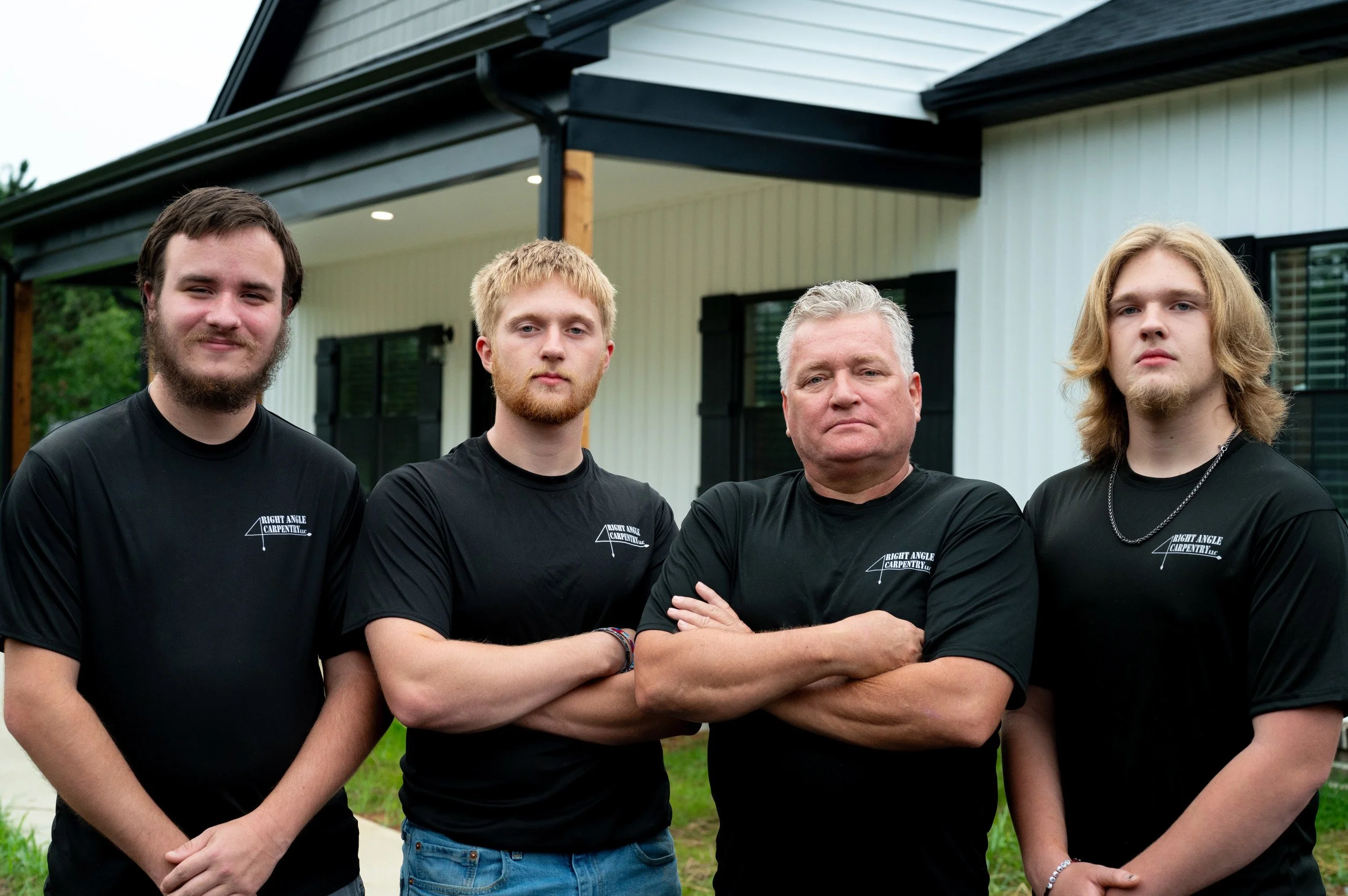 Group of four men standing outdoors in front of a house, wearing black t-shirts with a small logo on the left chest that reads 'Right Angle Carpentry'.