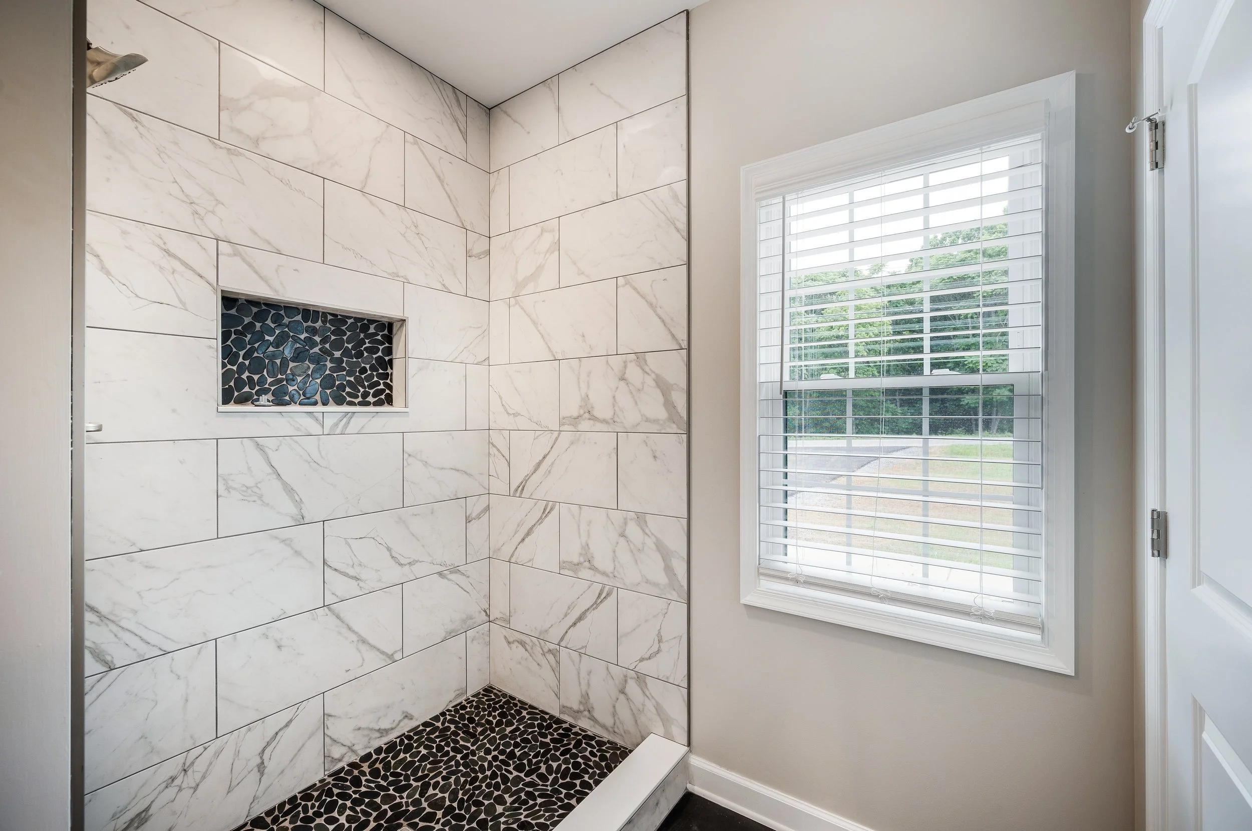 Bathroom shower with white marble tiles, black pebble floor and a built-in niche with black pebbles, next to a window with white blinds.
