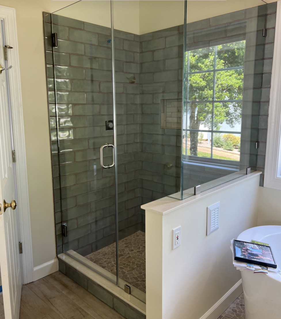 Bathroom with glass-enclosed walk-in shower featuring greenish-gray subway tiles, a window with a view of trees outside, and a small built-in shelf inside the shower. Next to the shower is a wall with a vent and a switch, and a bathtub with a magazine and some papers on the edge.