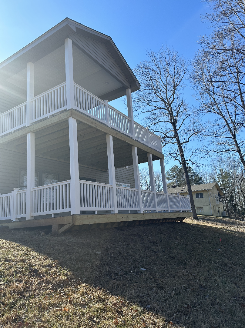 A two-story house with large balconies and white railings on a grassy hill, with leafless trees and a blue sky in the background.