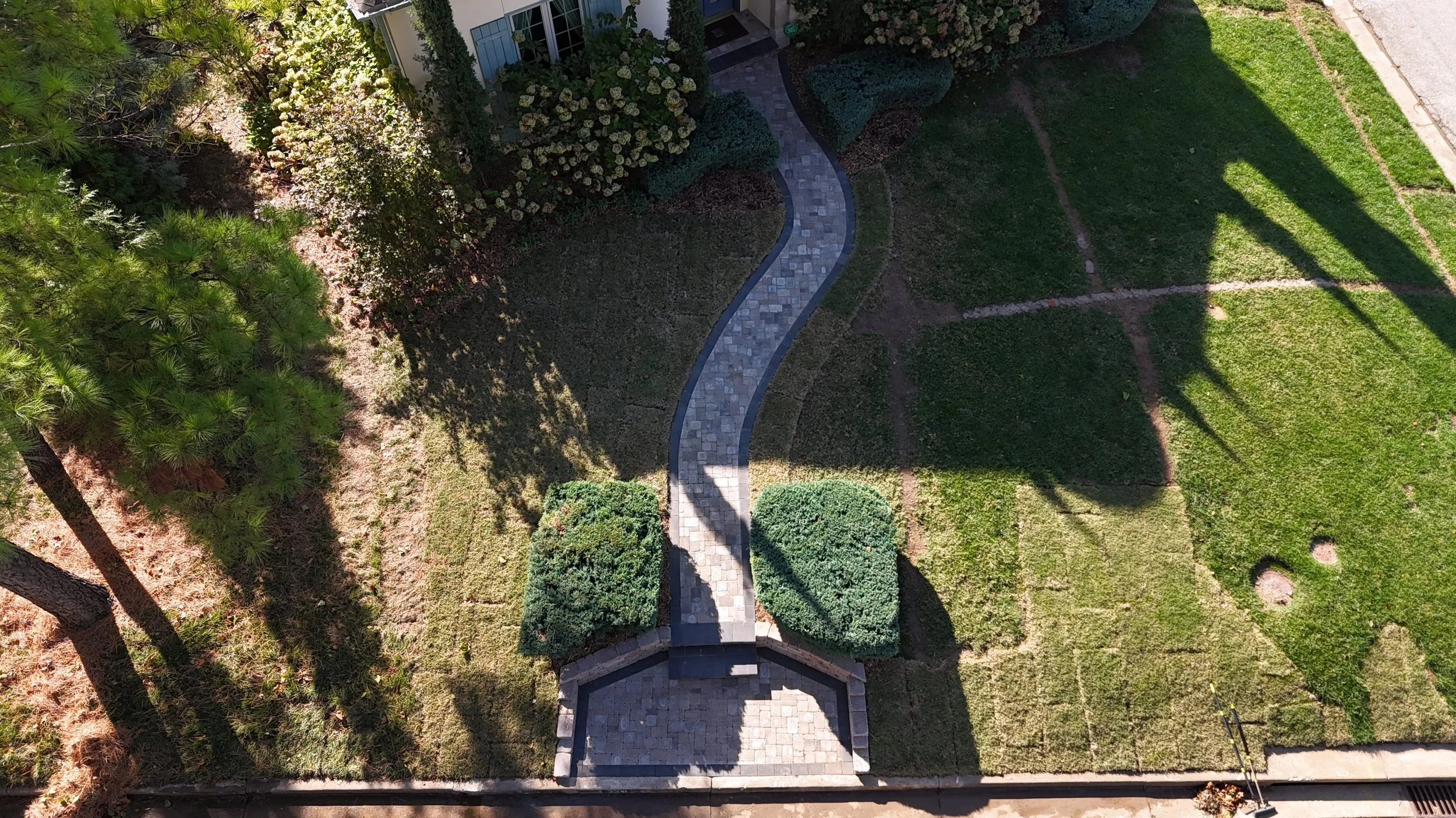 Aerial view of a landscaped yard with a curved stone pathway, green grass, bushes, and trees, with shadows cast across the garden.