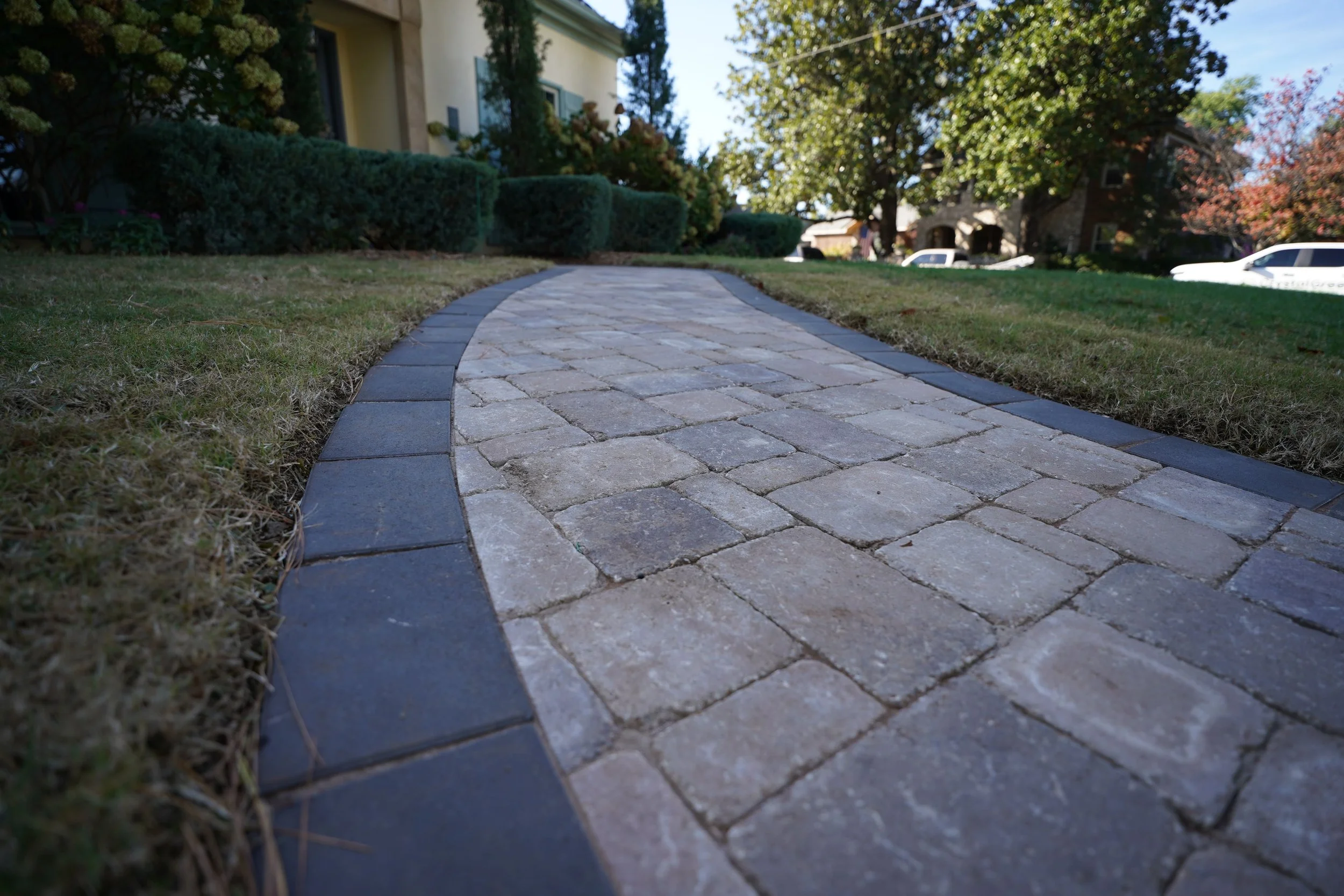 A curved brick walkway leading to a house, bordered by grass and shrubs, with trees and cars in the background.