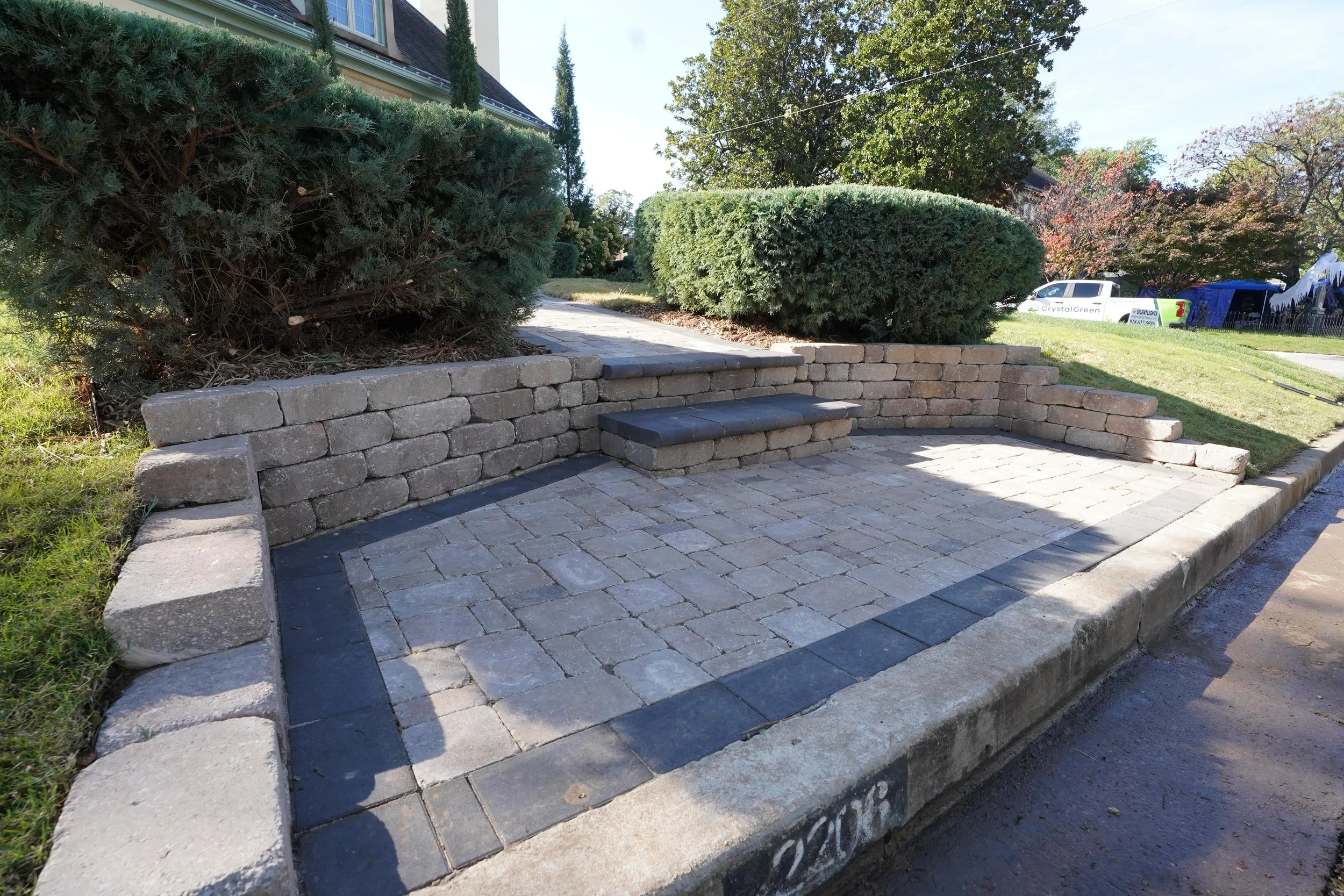 A small paved patio area with brick and stone steps, surrounded by brick retaining walls, green shrubs, and a lawn, with houses and trees in the background.