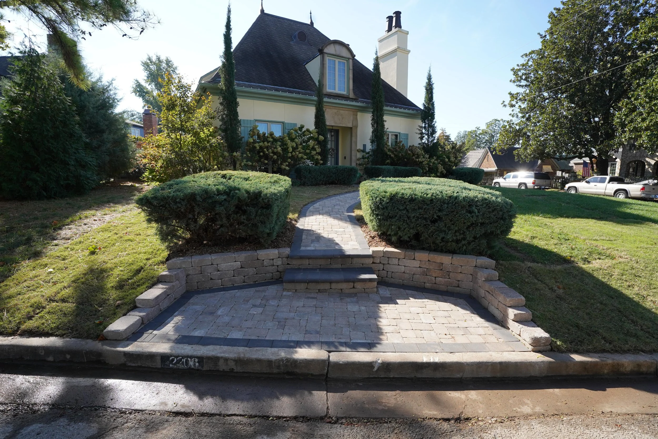 A curved brick and stone walkway leading to the front door of a house, surrounded by trimmed bushes and trees.