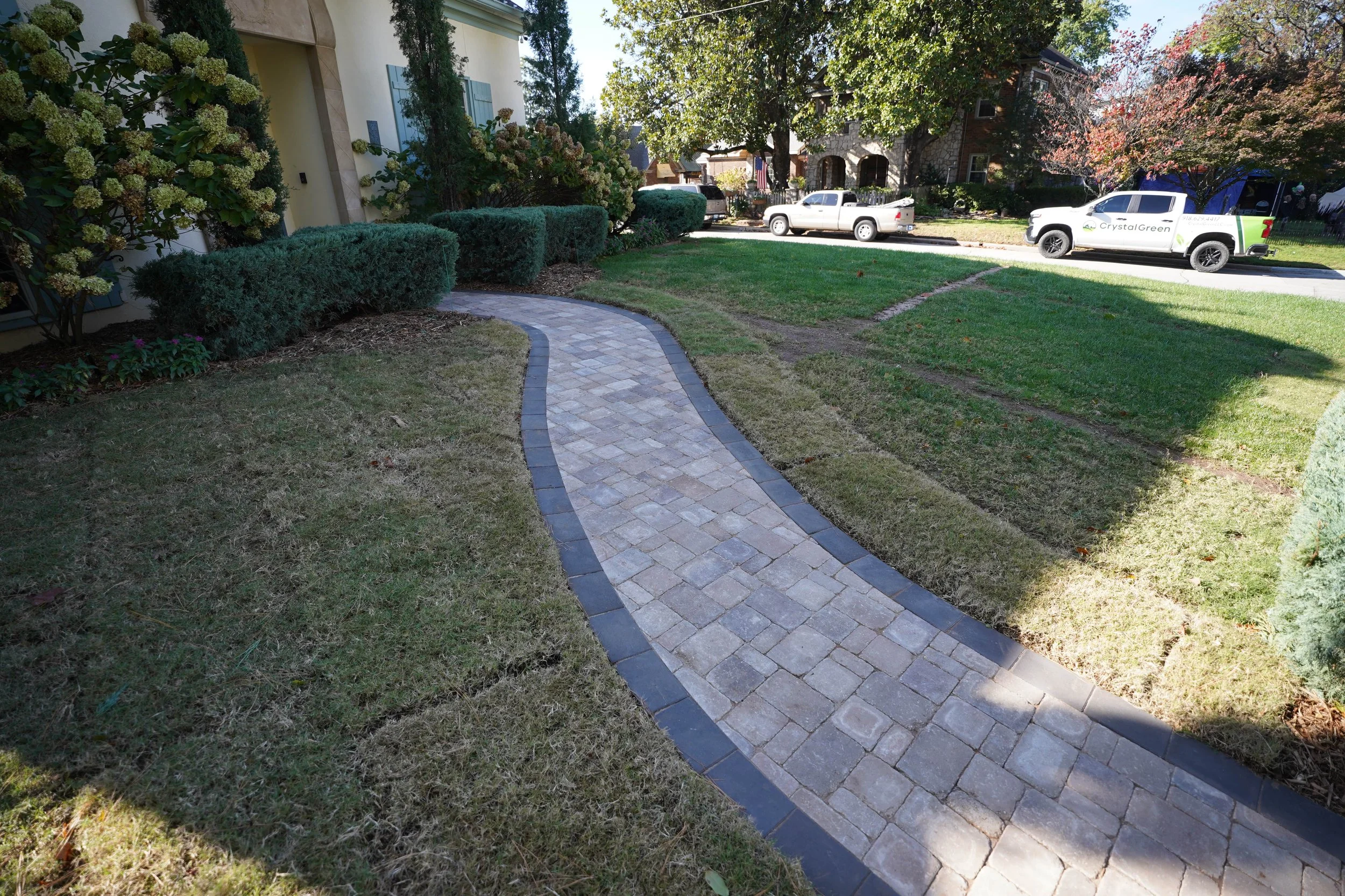 A curved stone walkway in a front yard with trimmed bushes and trees, adjacent to a lawn and residential houses with cars parked along the street.