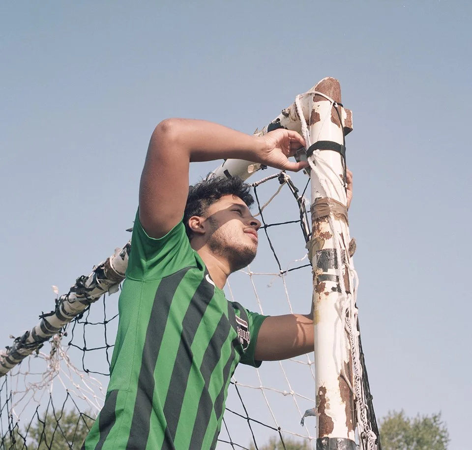 Man in green and black striped jersey adjusting a soccer goal net outdoors under clear sky.