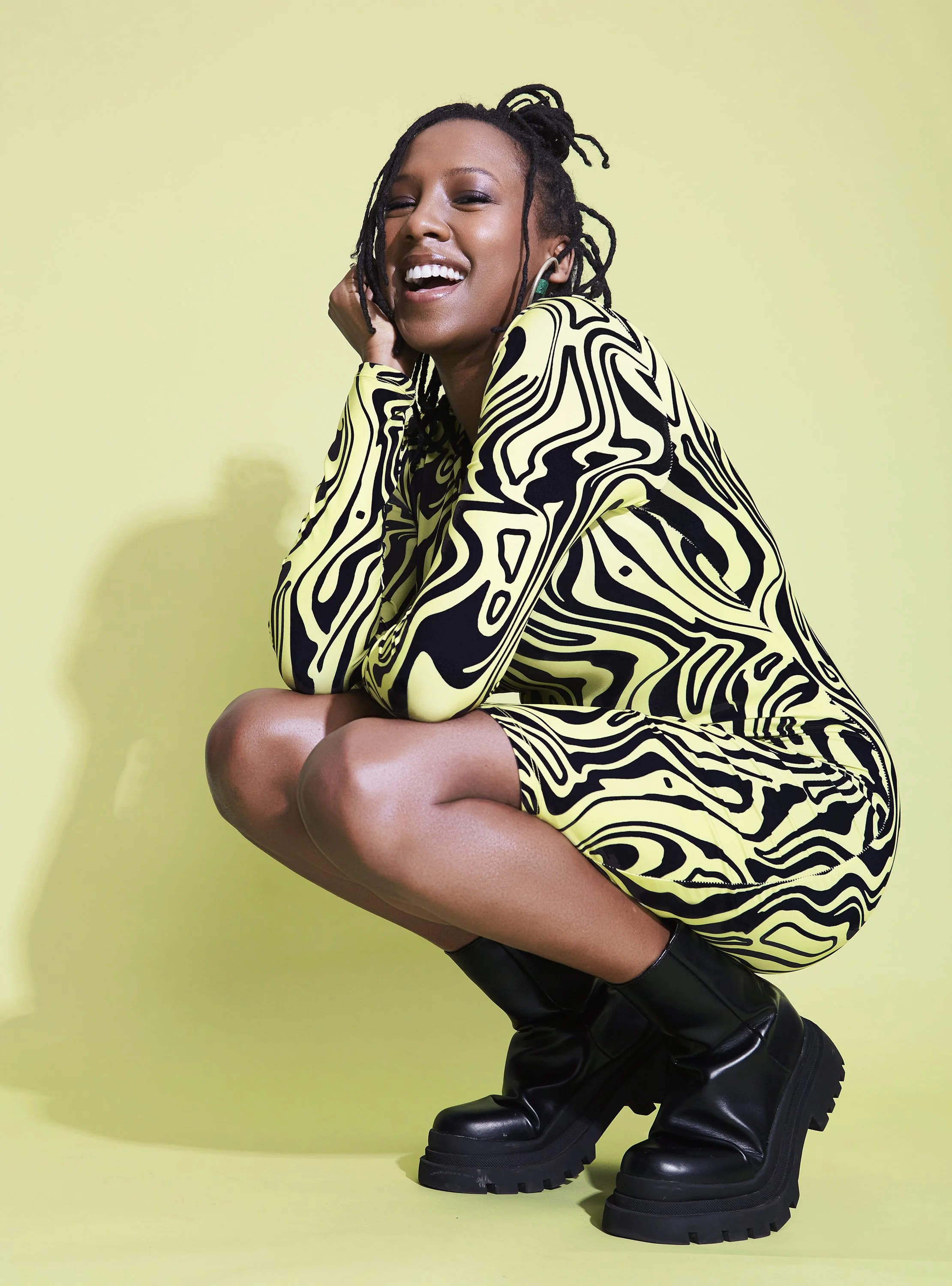 Person wearing a black and yellow patterned dress, black boots, crouching and smiling on a yellow background.