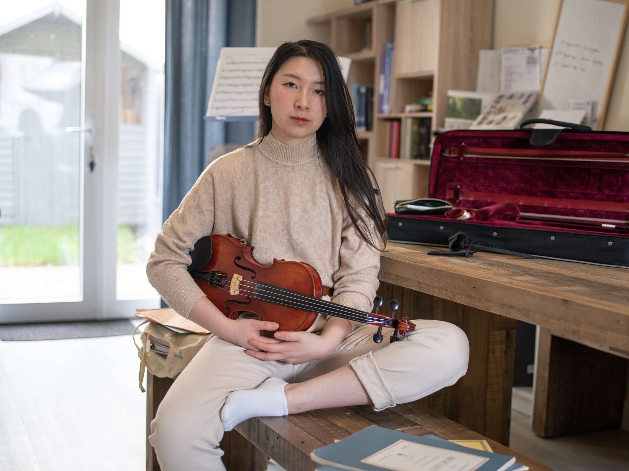 Person holding a violin seated on a wooden bench with an open violin case, music stand, and books in the background.